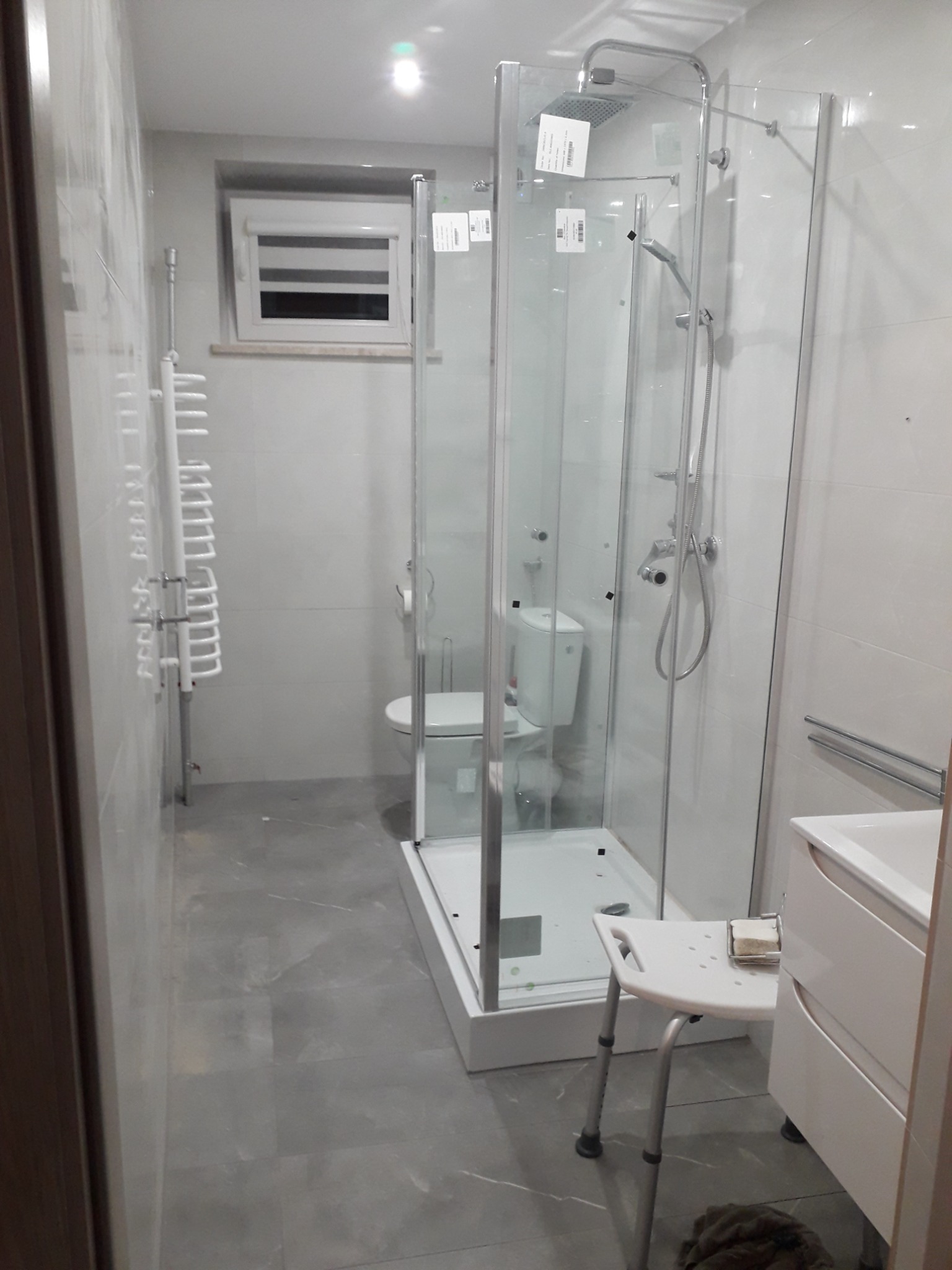 Nowoczesna łazienka z kabiną prysznicową typu walk-in, białymi płytkami na ścianach i szarymi płytkami podłogowymi. Widoczny biały grzejnik drabinkowy, toaleta oraz krzesło prysznicowe z mydłem.