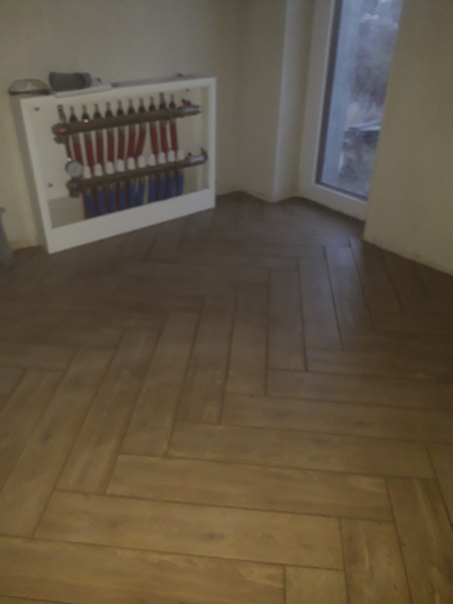 Ułożona w jodełkę brązowa podłoga z drewnopodobnych paneli, przy ścianie biała szafka z instalacją grzewczą z czerwonymi i niebieskimi rurkami, widoczny fragment okna.