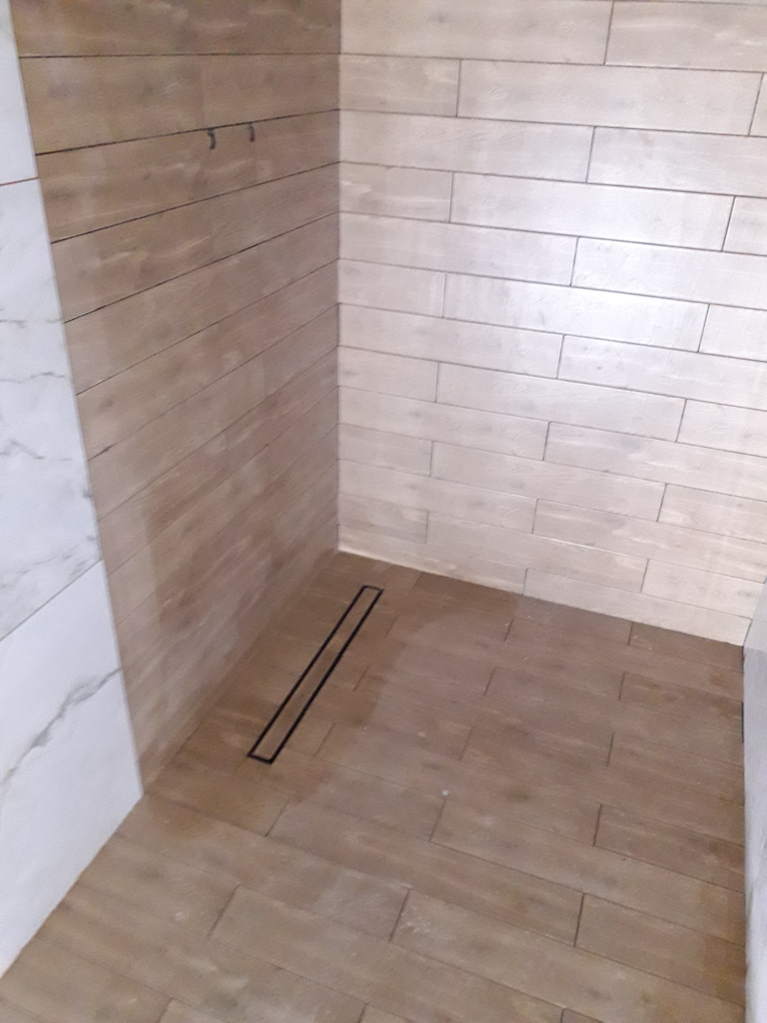 Ukończona kabina prysznicowa z odpływem liniowym, wyłożona płytkami imitującymi drewno, z białymi płytkami marmurkowymi po lewej stronie.