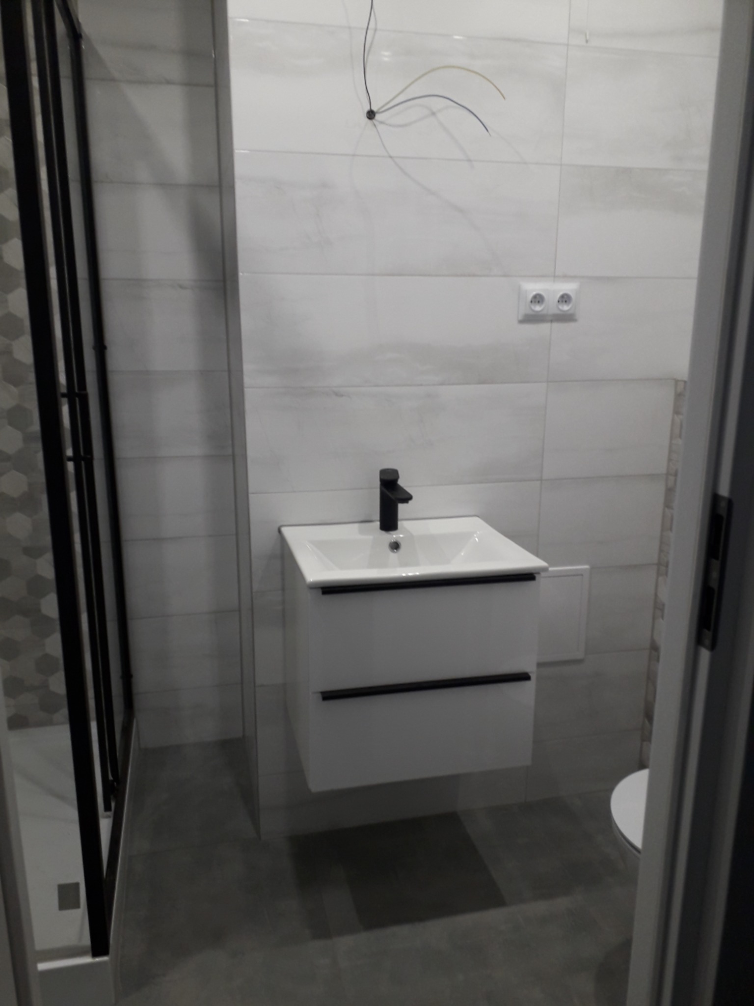 Nowoczesna łazienka z białą szafką pod umywalkę z czarnymi uchwytami i czarną baterią, szara podłoga i płytki, częściowo widoczna kabina prysznicowa z czarnymi profilami i wystające kable z sufitu.
