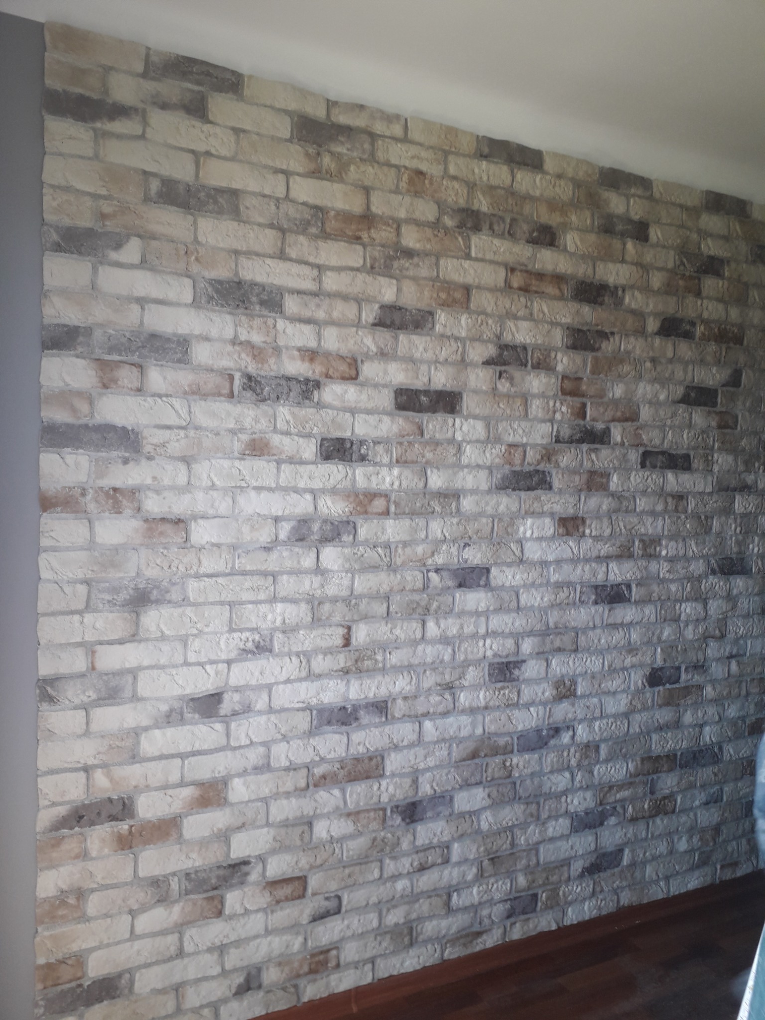 Ściana z dekoracyjnej cegły w odcieniach bieli, szarości i brązu, z ciemną drewnianą podłogą u dołu kadru.