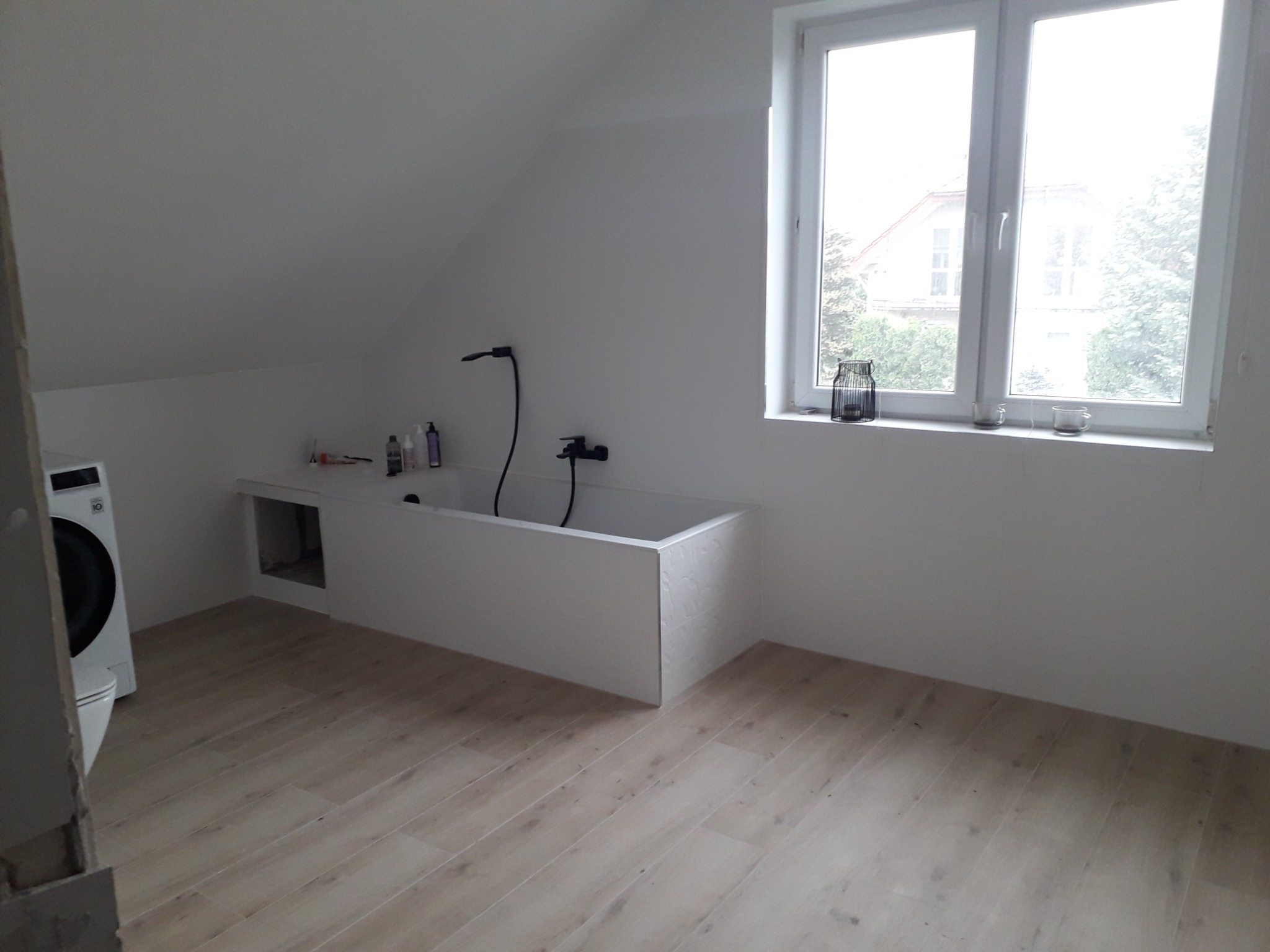 Nowoczesna łazienka na poddaszu z białą wanną w zabudowie, czarną armaturą i drewnopodobną podłogą; pralka widoczna z lewej strony.