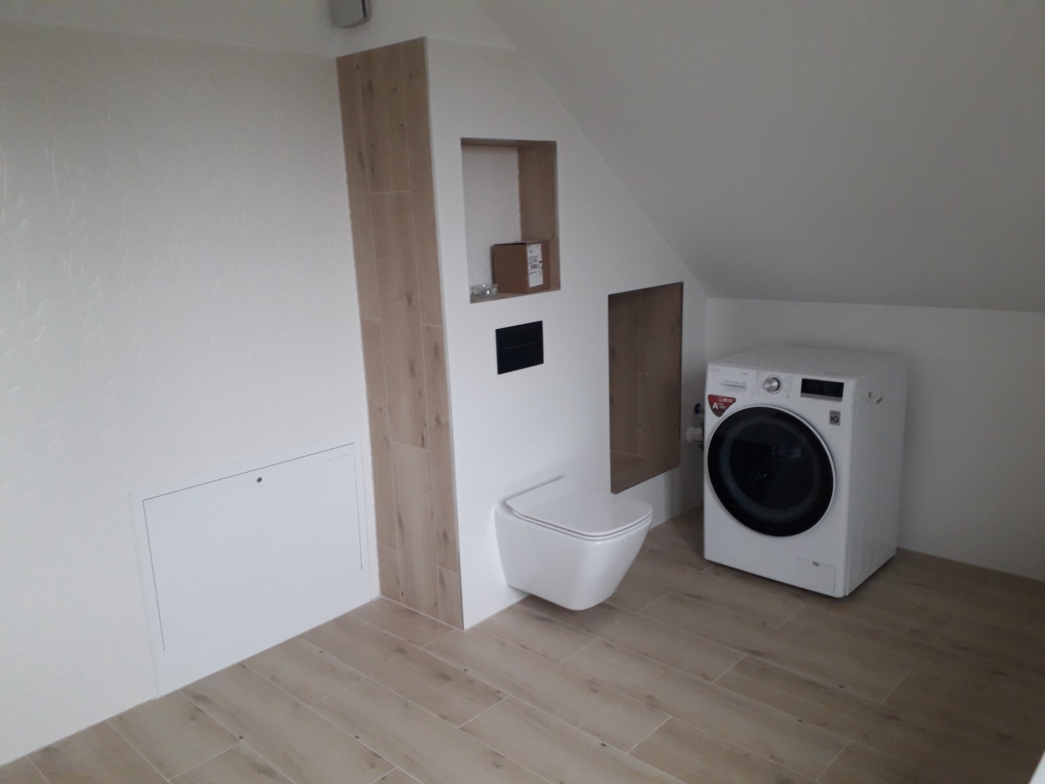 Ukończona łazienka na poddaszu z podwieszaną toaletą, drewnopodobnymi płytkami podłogowymi i pralką LG.