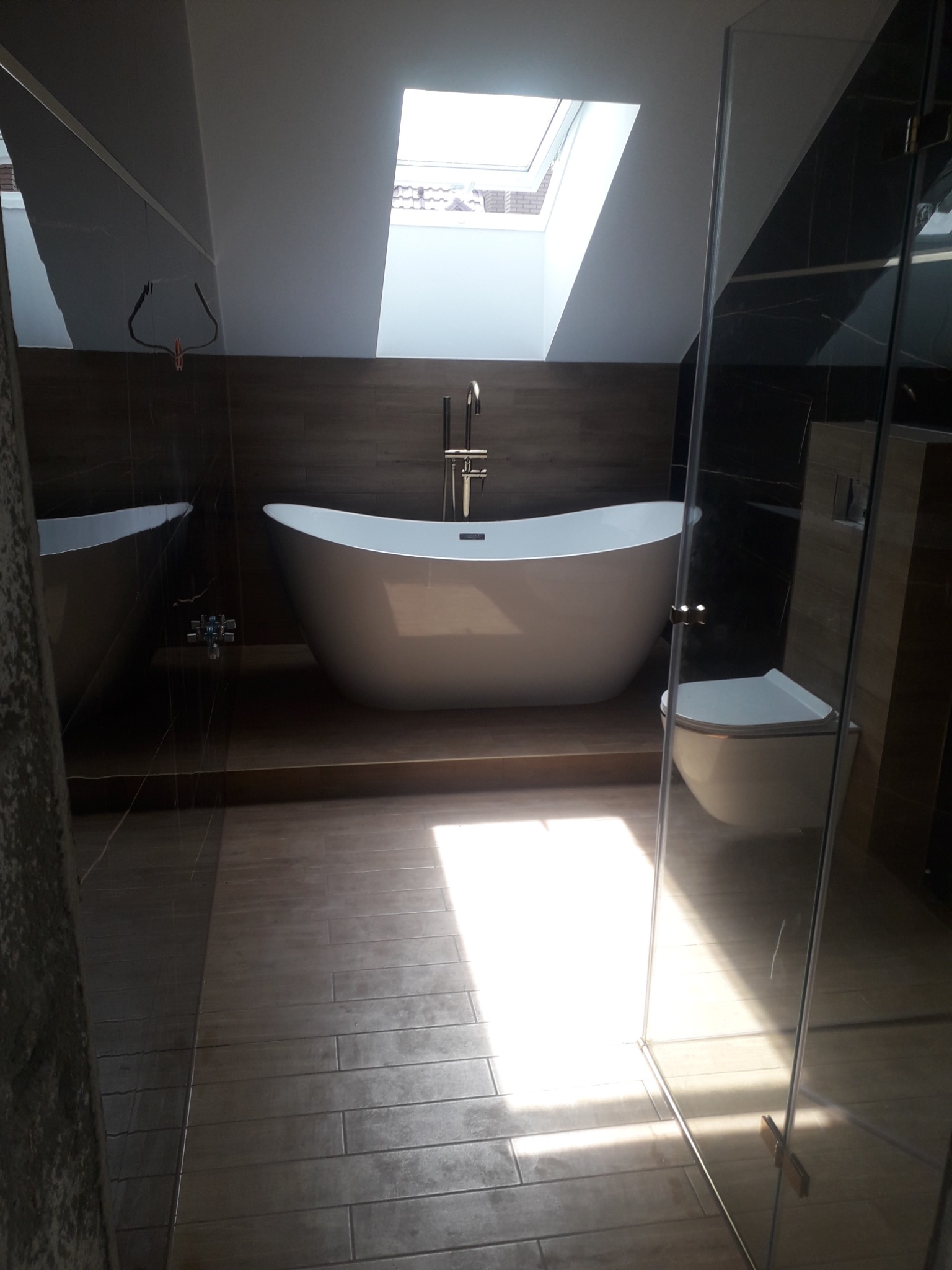 Nowoczesna łazienka na poddaszu z wolnostojącą wanną, toaletą i kabiną prysznicową, oświetlona światłem wpadającym przez okno dachowe.