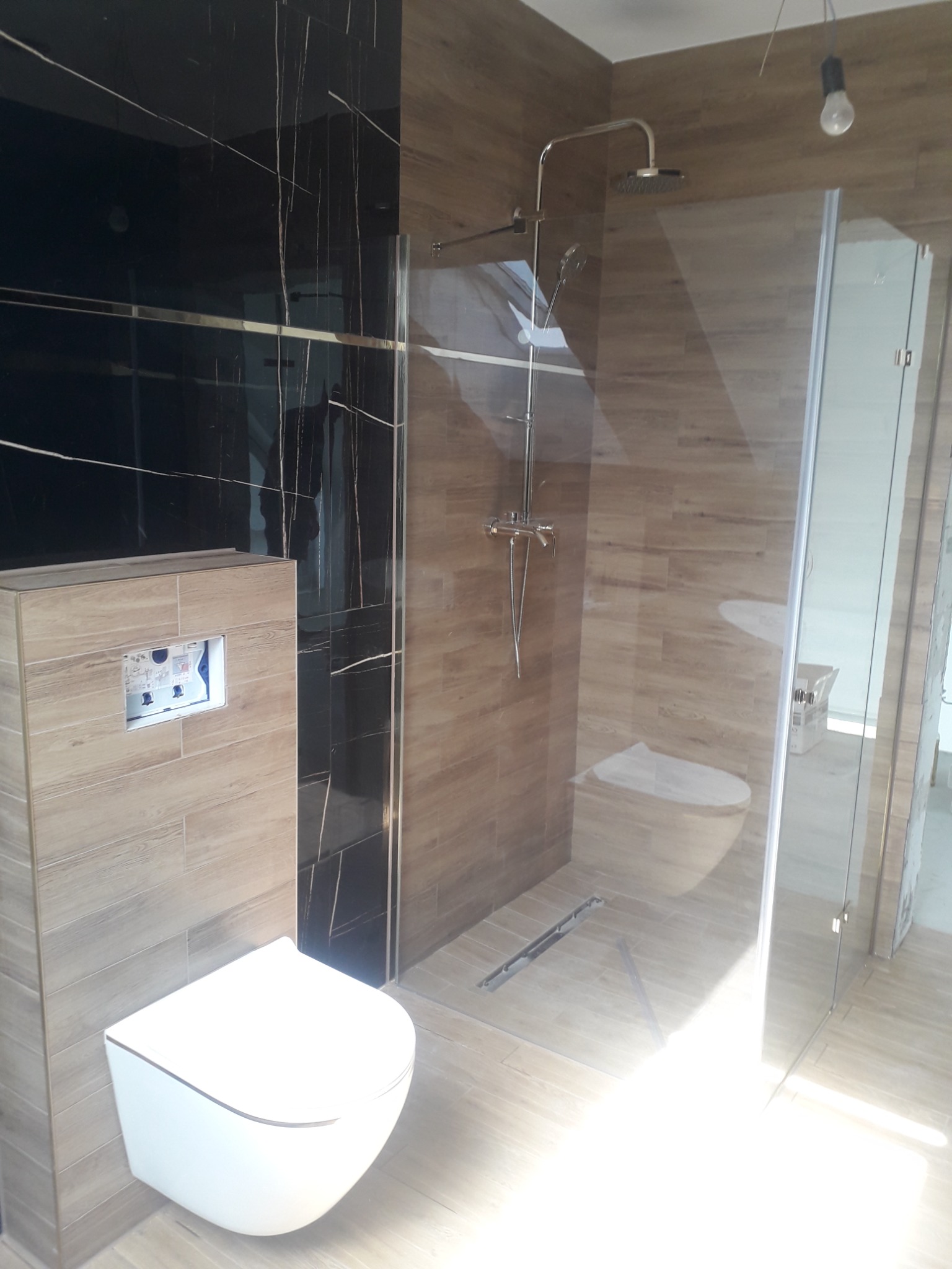 Nowoczesna łazienka po remoncie w Małogoszczu: wisząca toaleta, kabina prysznicowa ze szklanymi drzwiami i deszczownicą, płytki imitujące drewno oraz czarne płytki z białymi żyłami.