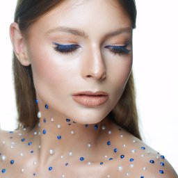 Kate_marko - Portret kobiety z delikatnym makijażem oczu w odcieniach brązu i niebieskiego, ozdobionej perłami i niebieskimi koralikami na dekolcie i ramionach, na jasnym tle.