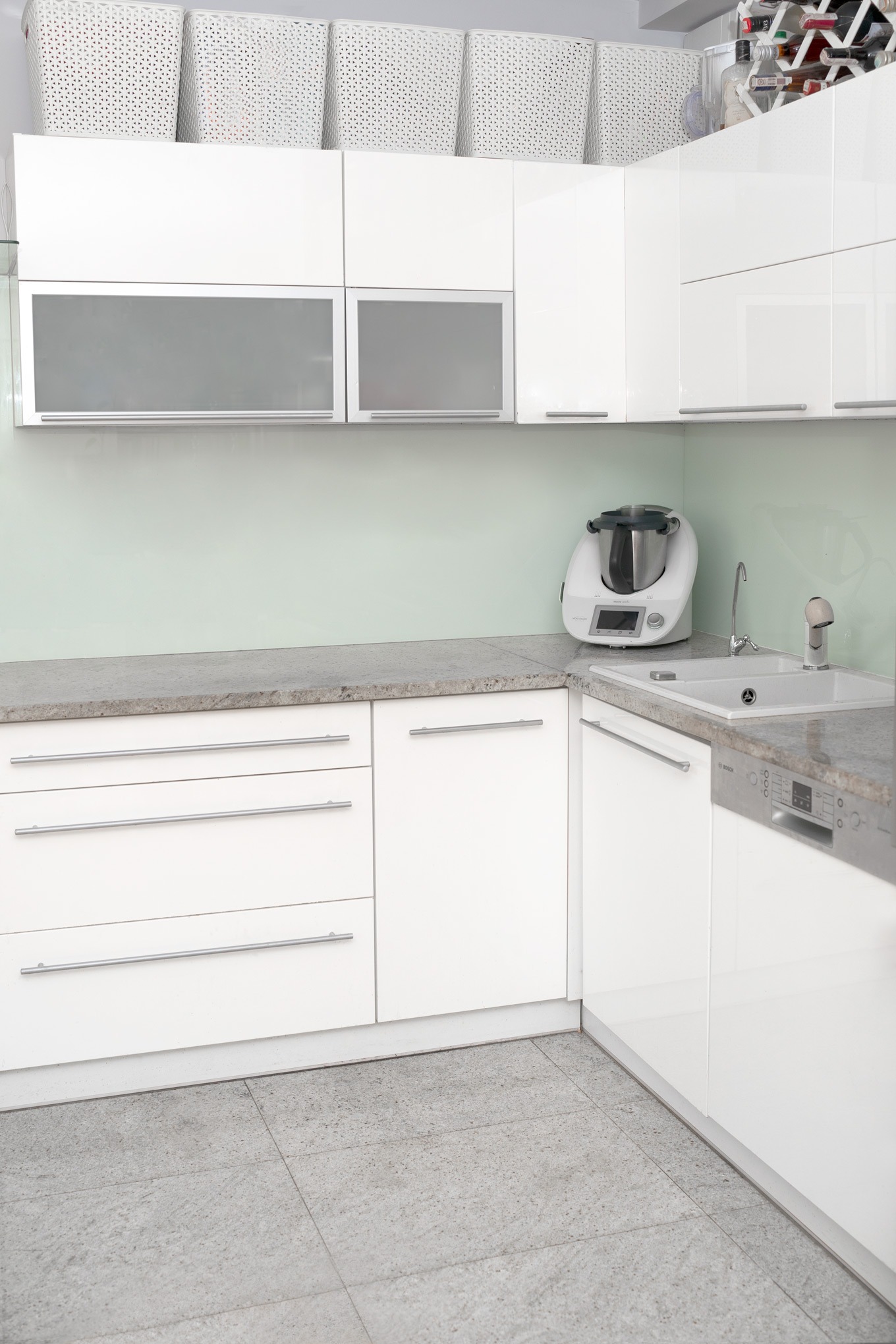 Jasna, nowoczesna kuchnia z białymi szafkami, szarym blatem i podłogą z płytek. Na blacie stoi biały robot kuchenny Thermomix, a nad szafkami wiszą białe, ażurowe kosze.