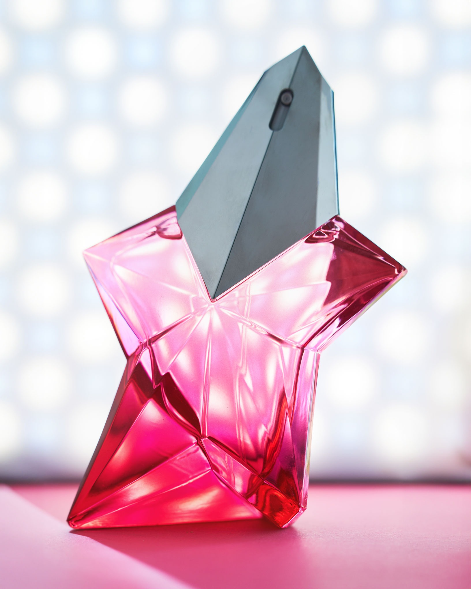 Błyszczący, różowy flakon perfum w kształcie gwiazdy z szarym, geometrycznym korkiem na tle rozmytego, jasnego tła z okrągłymi refleksami.