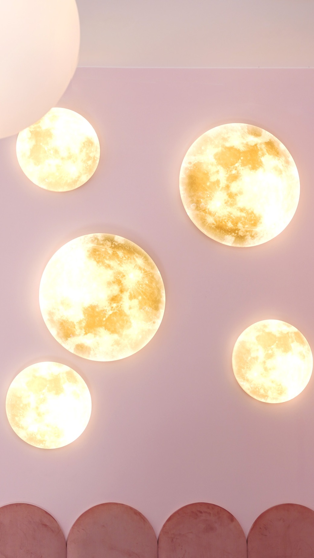 Różowa ściana z pięcioma okrągłymi lampami przypominającymi księżyc w pełni, z miękkim światłem i pluszowym panelem dekoracyjnym u dołu.