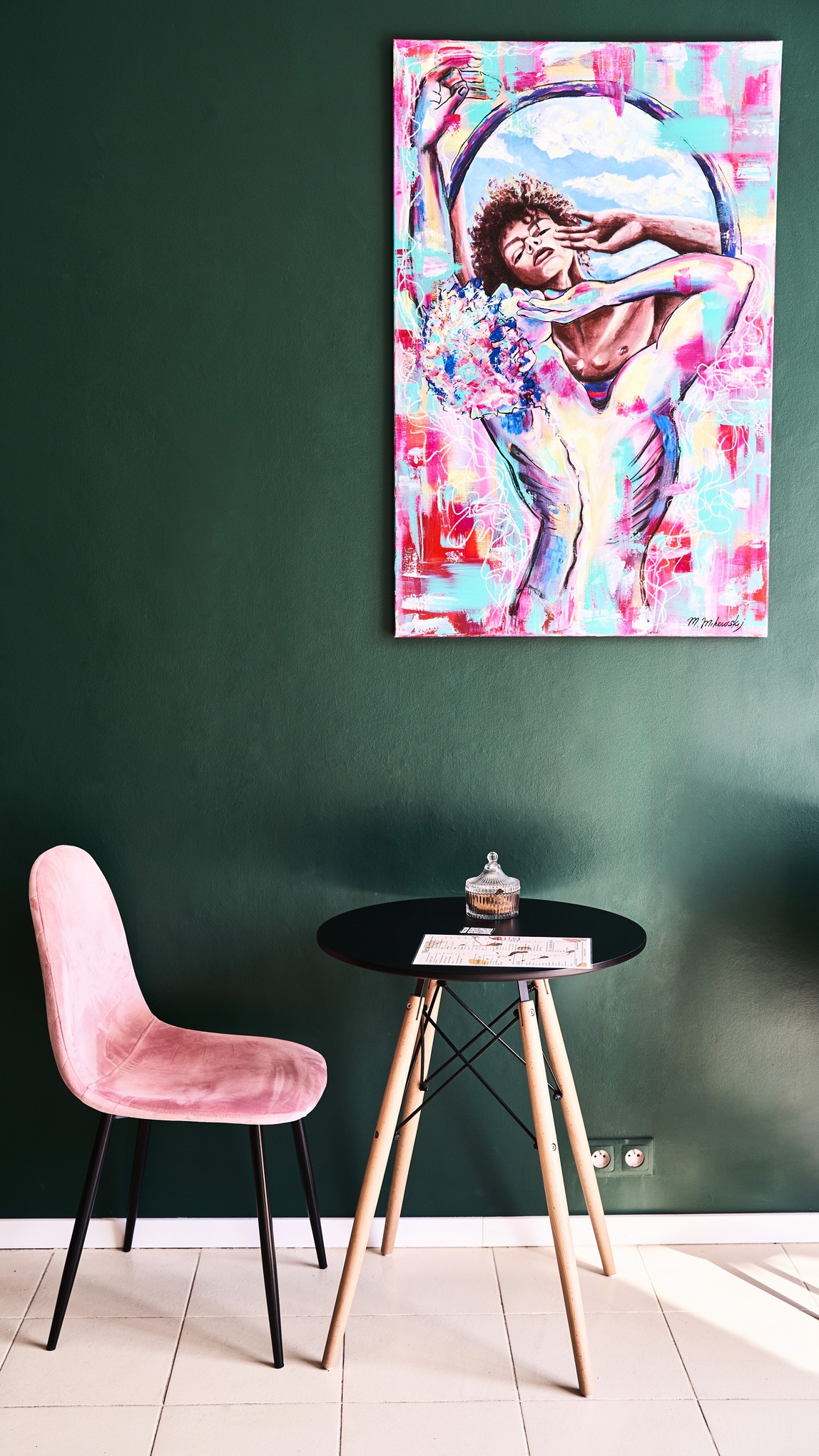 Wnętrze kawiarni z ciemnozieloną ścianą, na której wisi abstrakcyjny obraz przedstawiający postać, obok stoi różowe krzesło i mały czarny stolik z menu i szklanym pojemnikiem.