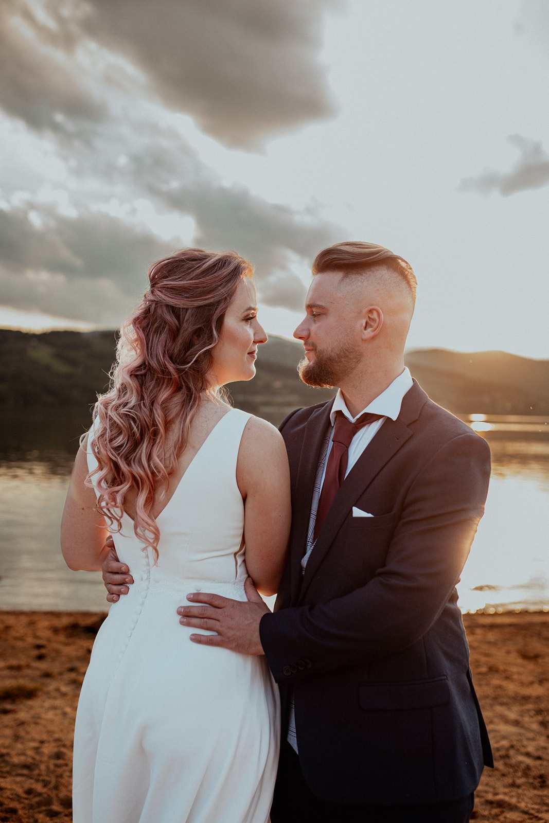 Para młoda w objęciach na tle jeziora i zachodzącego słońca, panna młoda z różowymi włosami i suknią z odkrytymi plecami, pan młody w granatowym garniturze i bordowym krawacie.