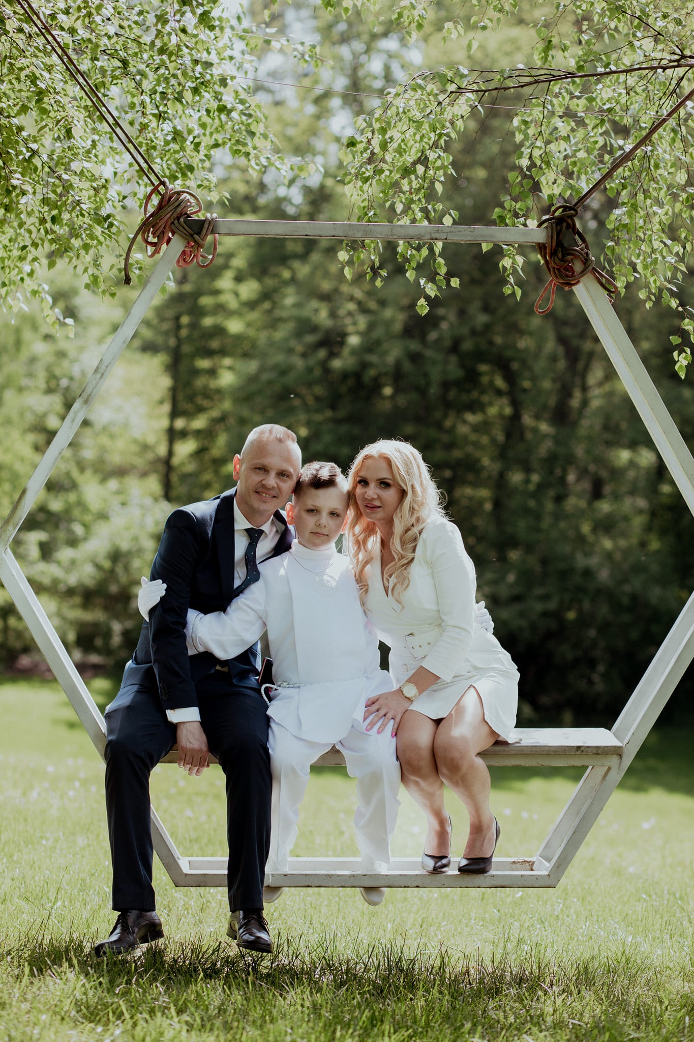 Rodzinne zdjęcie w plenerze: ojciec w granatowym garniturze obejmuje syna ubranego w biały strój komunijny, obok matka w białej sukience, wszyscy siedzą na metalowej huśtawce w kształcie...