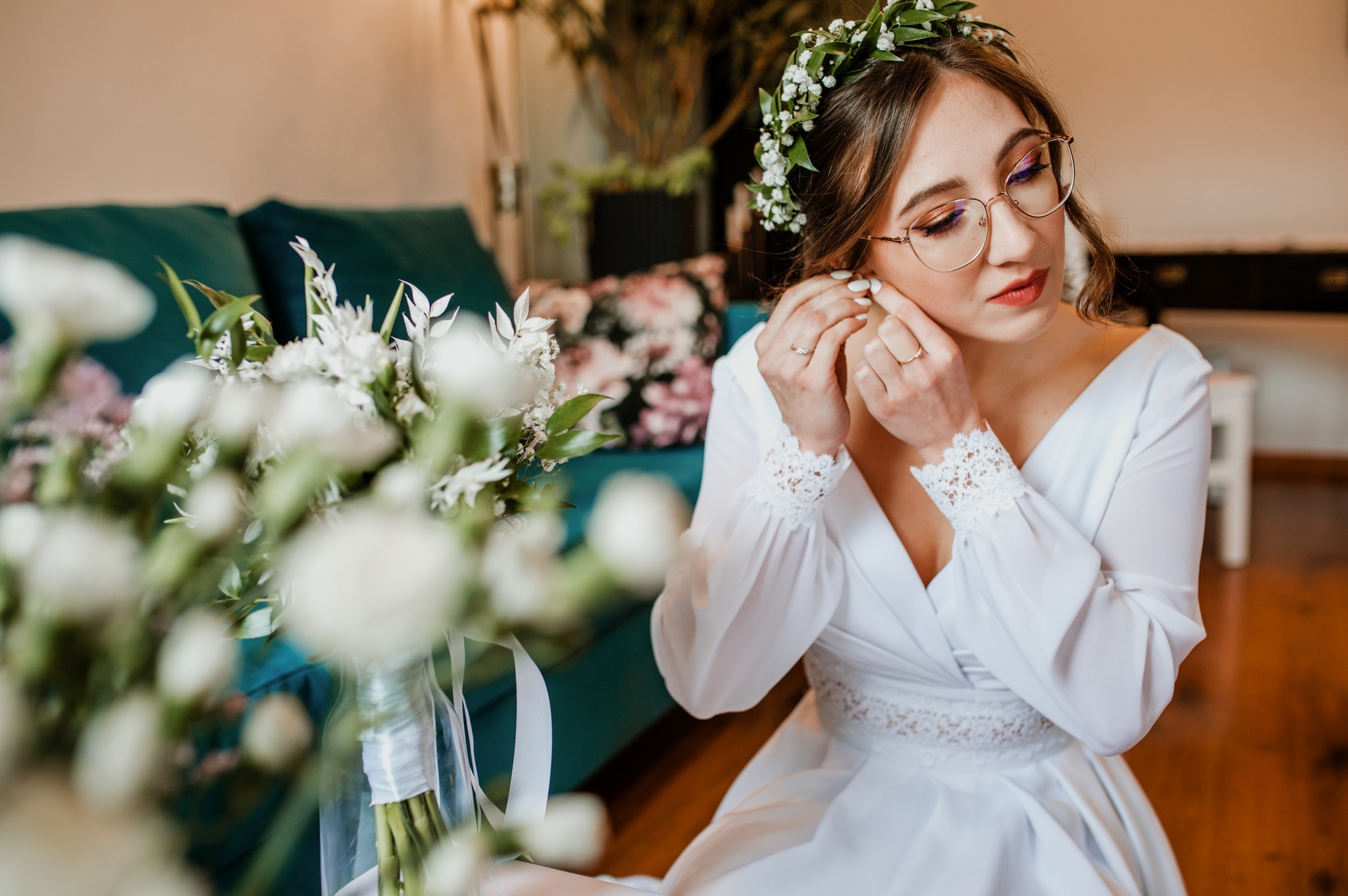 Panna młoda w białej sukni i wianku z drobnych kwiatów zakłada kolczyk, obok bukiet ślubny z białych kwiatów, widoczne okulary w złotej oprawce.