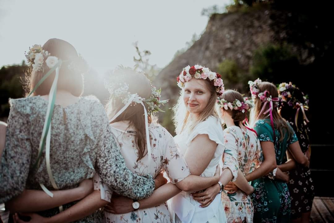 Grupa kobiet w wiankach obejmuje się na tle skalistego wzgórza w słoneczny dzień, jedna z kobiet patrzy w obiektyw.