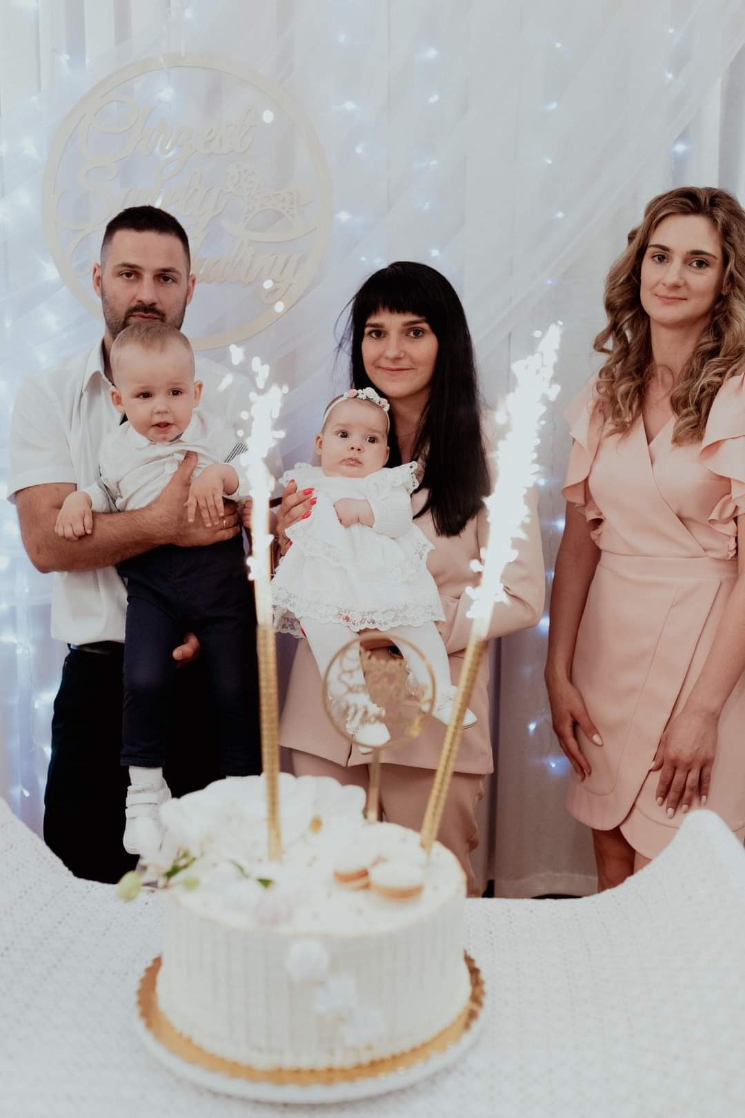 Rodzinne zdjęcie z dwójką małych dzieci, tortem z fontannami iskier i dekoracją z napisem 'Chrzest Święty' w tle, wykonane w jasnej kolorystyce.