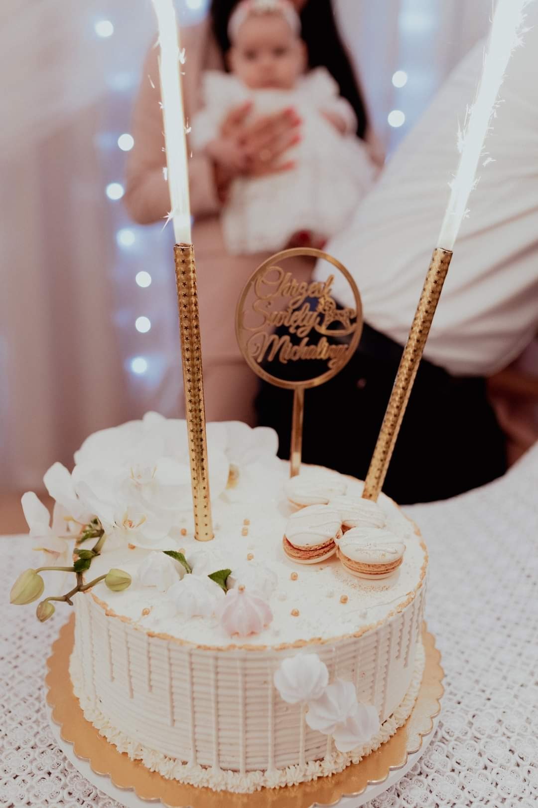 Tort z dekoracjami kwiatowymi, makaronikami i płonącymi fontannami, topper z napisem 'Chrzest Święty Michała', w tle osoba trzymająca dziecko w białej sukience.
