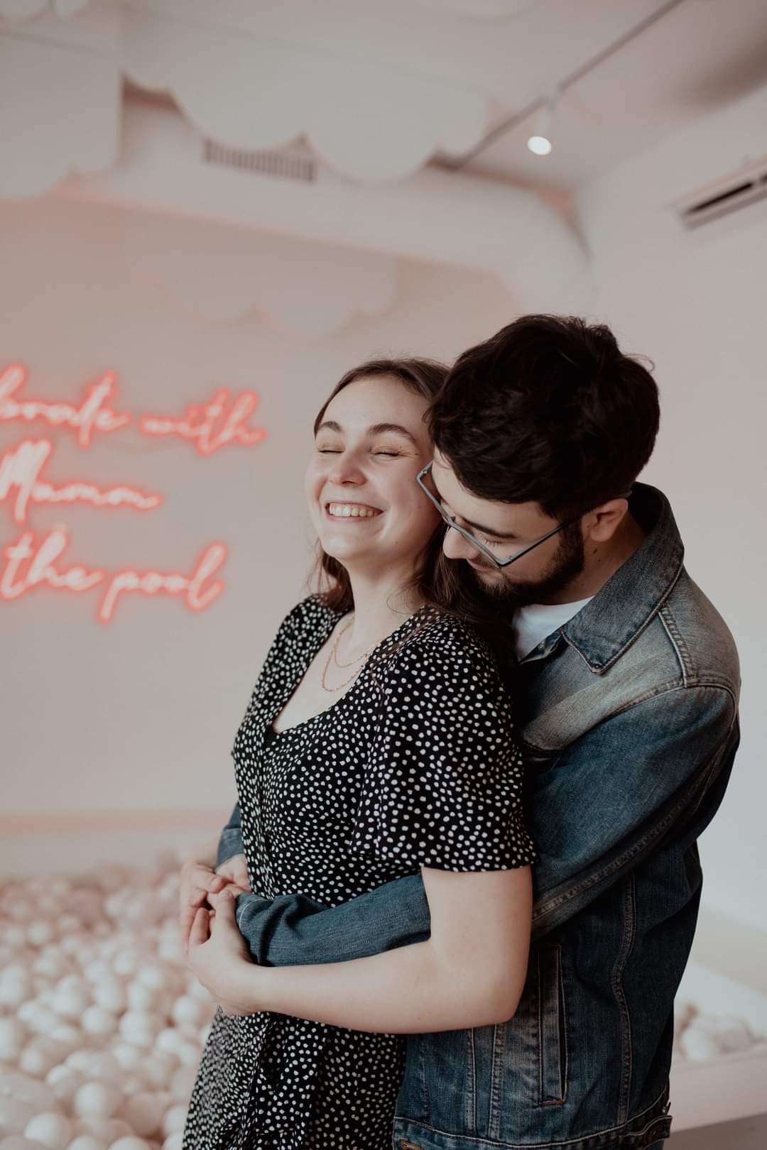 Uśmiechnięta para, kobieta w czarno-białej sukience i mężczyzna w jeansowej kurtce, obejmująca się na tle białych kul i neonowego napisu.