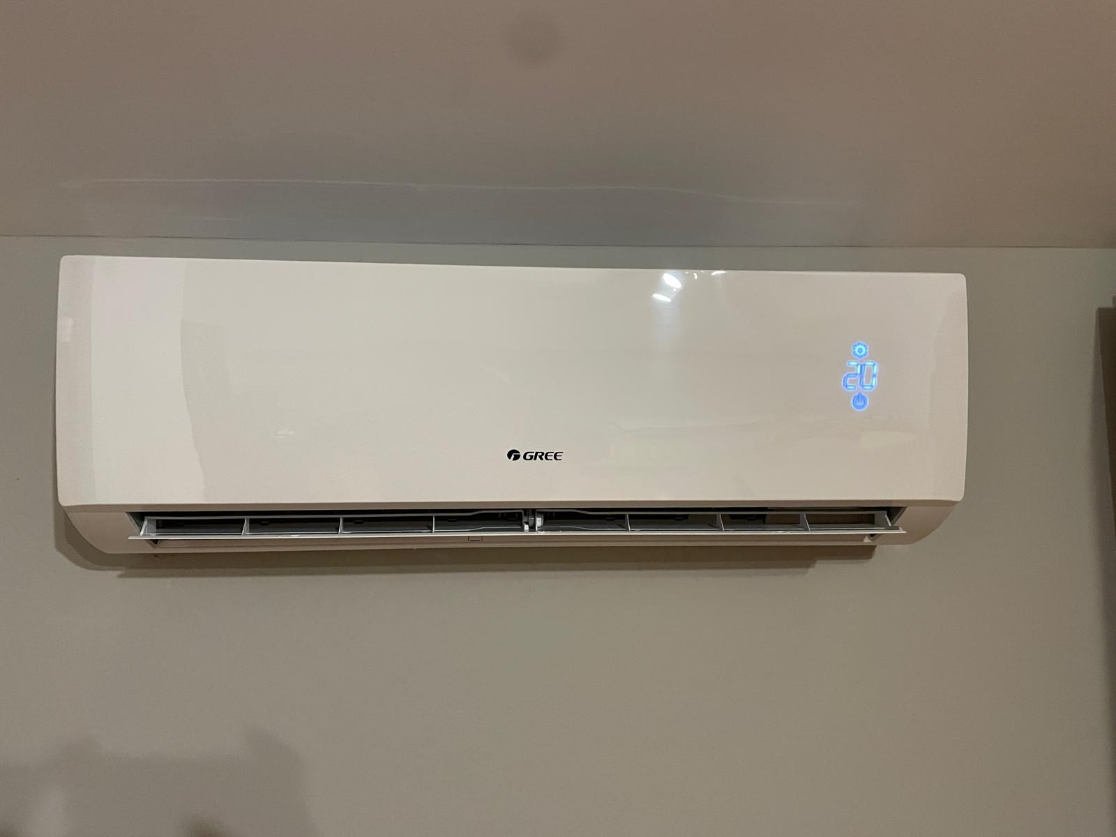 Biała klimatyzacja marki Gree zamontowana na szarej ścianie, wyświetlająca temperaturę 20 stopni Celsjusza.