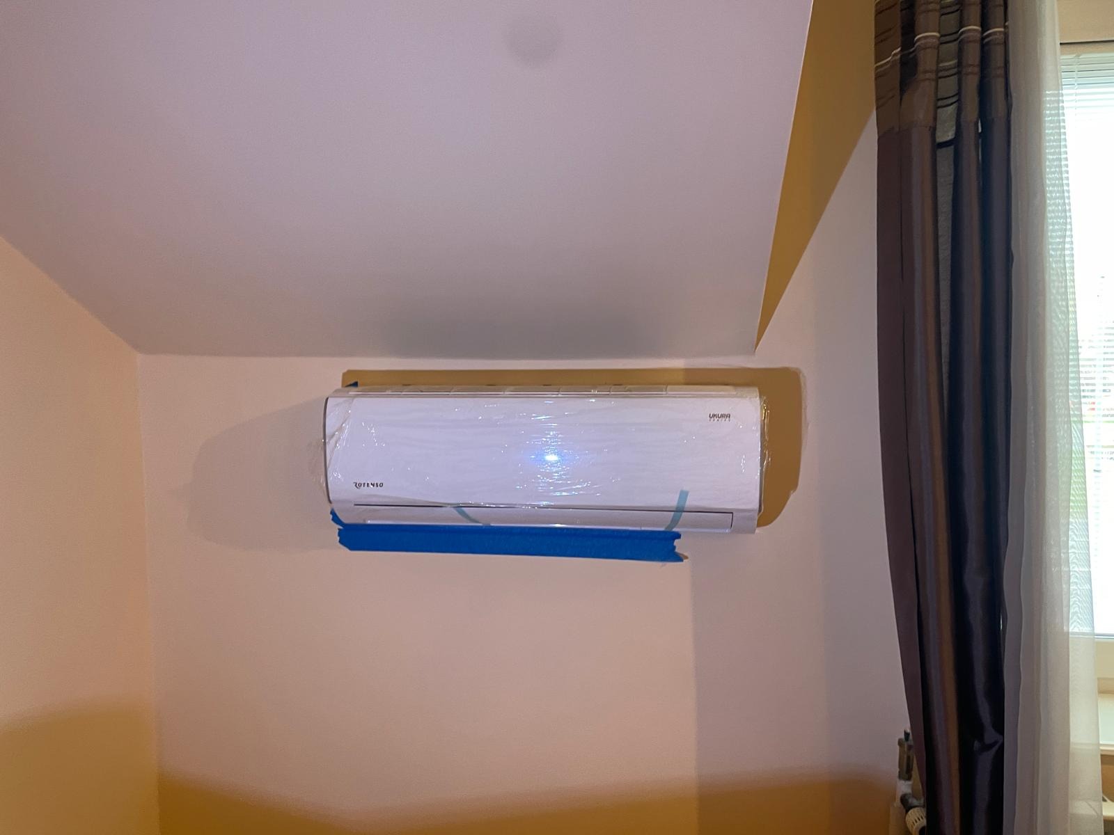 Biała jednostka wewnętrzna klimatyzatora marki Rotenso Ukura, jeszcze w folii ochronnej, zamontowana na beżowej ścianie w pomieszczeniu z brązowymi zasłonami i oknem.