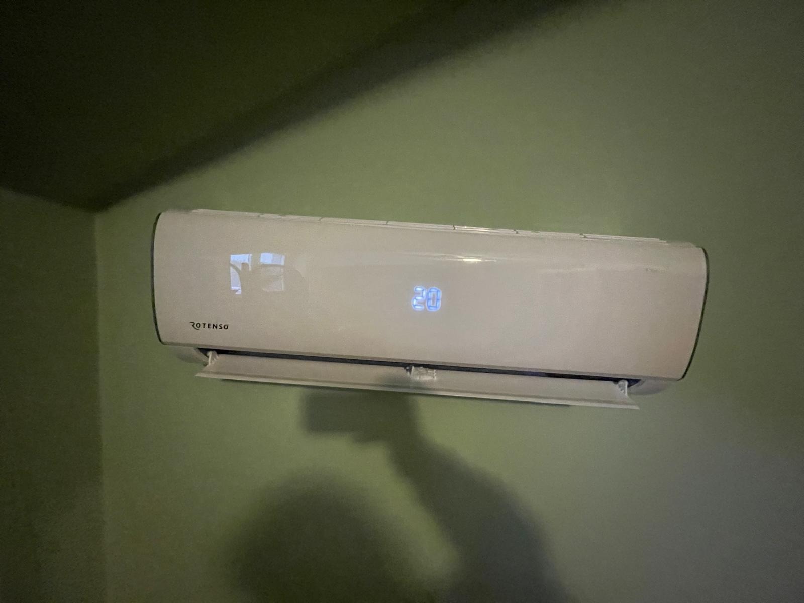 Biała jednostka wewnętrzna klimatyzatora Rotenso zamontowana na zielonej ścianie, wyświetlacz pokazuje 20 stopni Celsjusza, widoczny cień osoby robiącej zdjęcie.