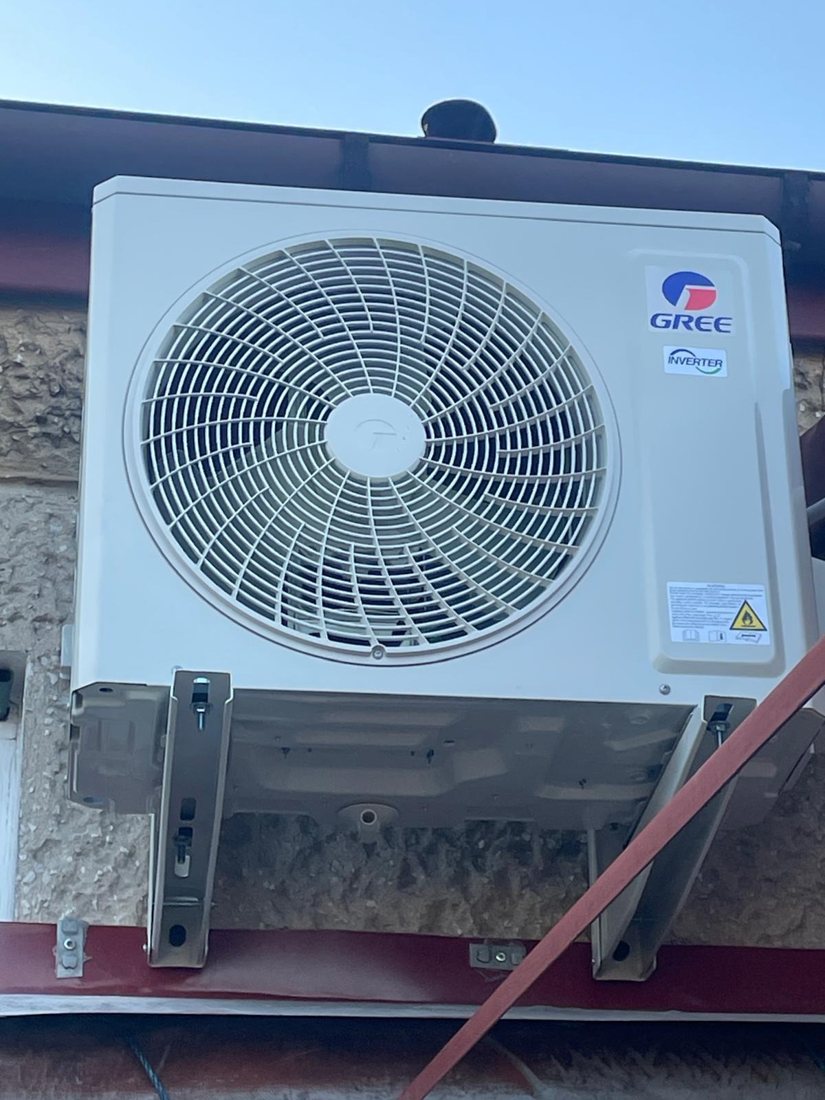 Zewnętrzna jednostka klimatyzacji GREE Inverter zamontowana na ścianie budynku z szorstką fakturą, widoczne metalowe wsporniki i fragment dachu.