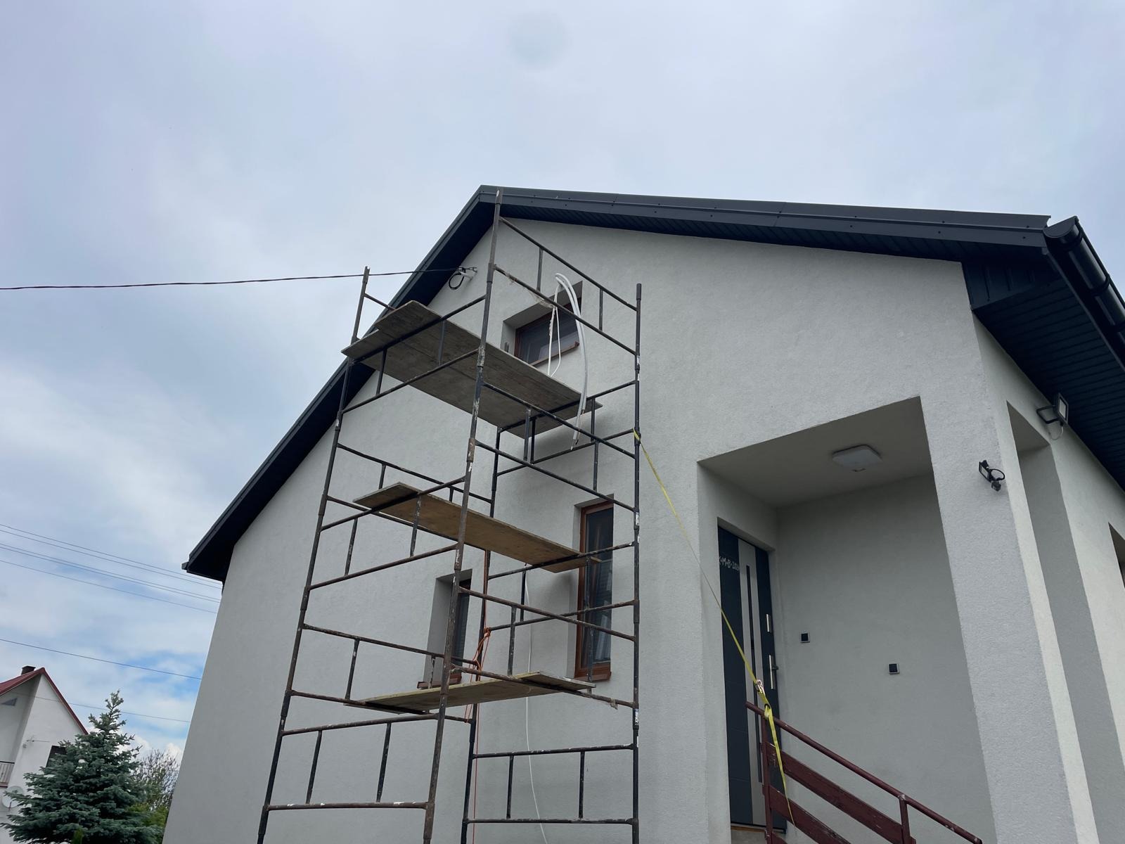 Elewacja domu jednorodzinnego w trakcie prac wykończeniowych, widoczne rusztowanie z drewnianymi podestami, szara elewacja i ciemna stolarka okienna oraz drzwiowa.