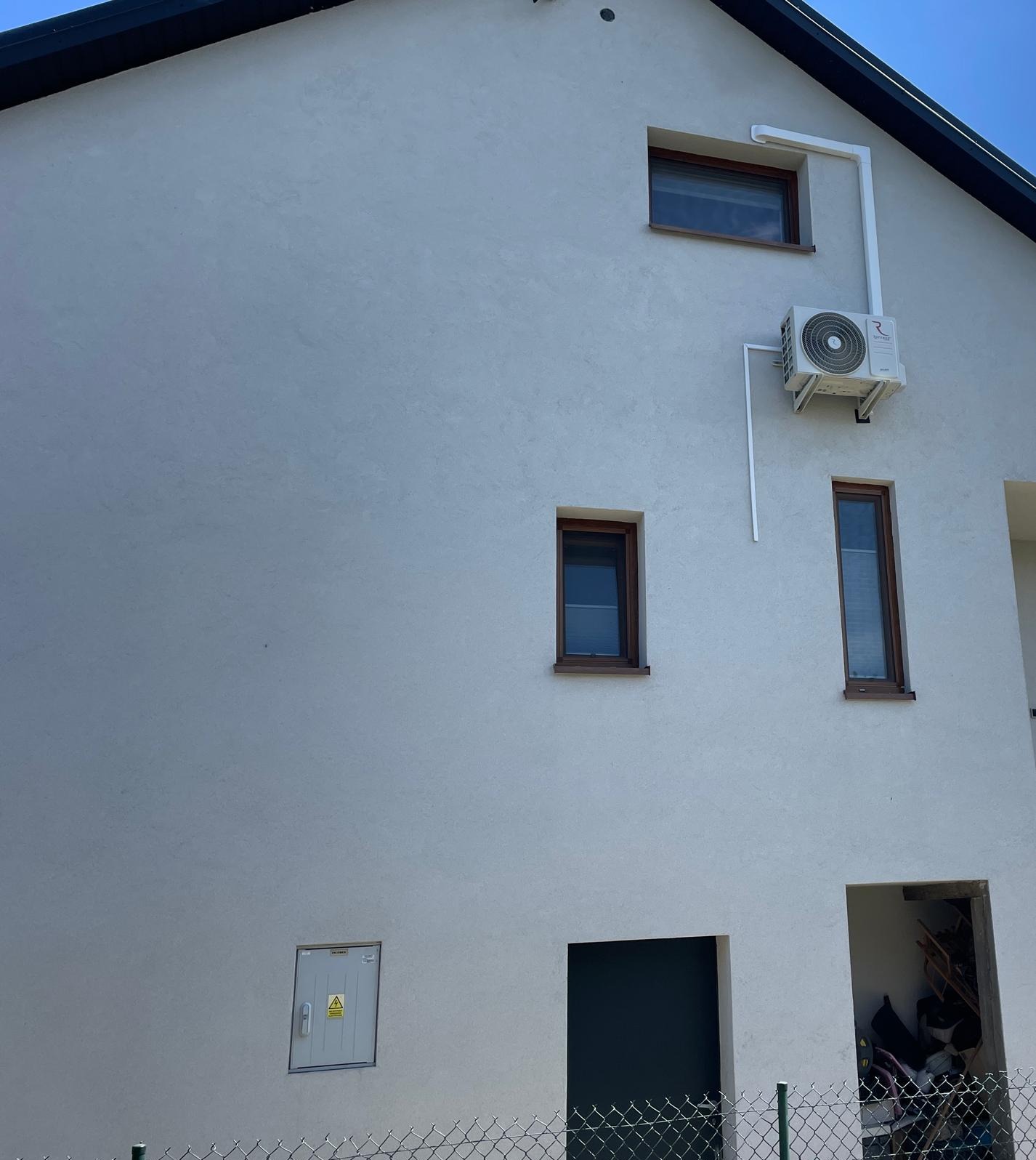 Biała ściana domu z zamontowaną jednostką zewnętrzną klimatyzacji, widoczne okna z brązowymi ramami, skrzynka elektryczna i fragment ogrodzenia z siatki.