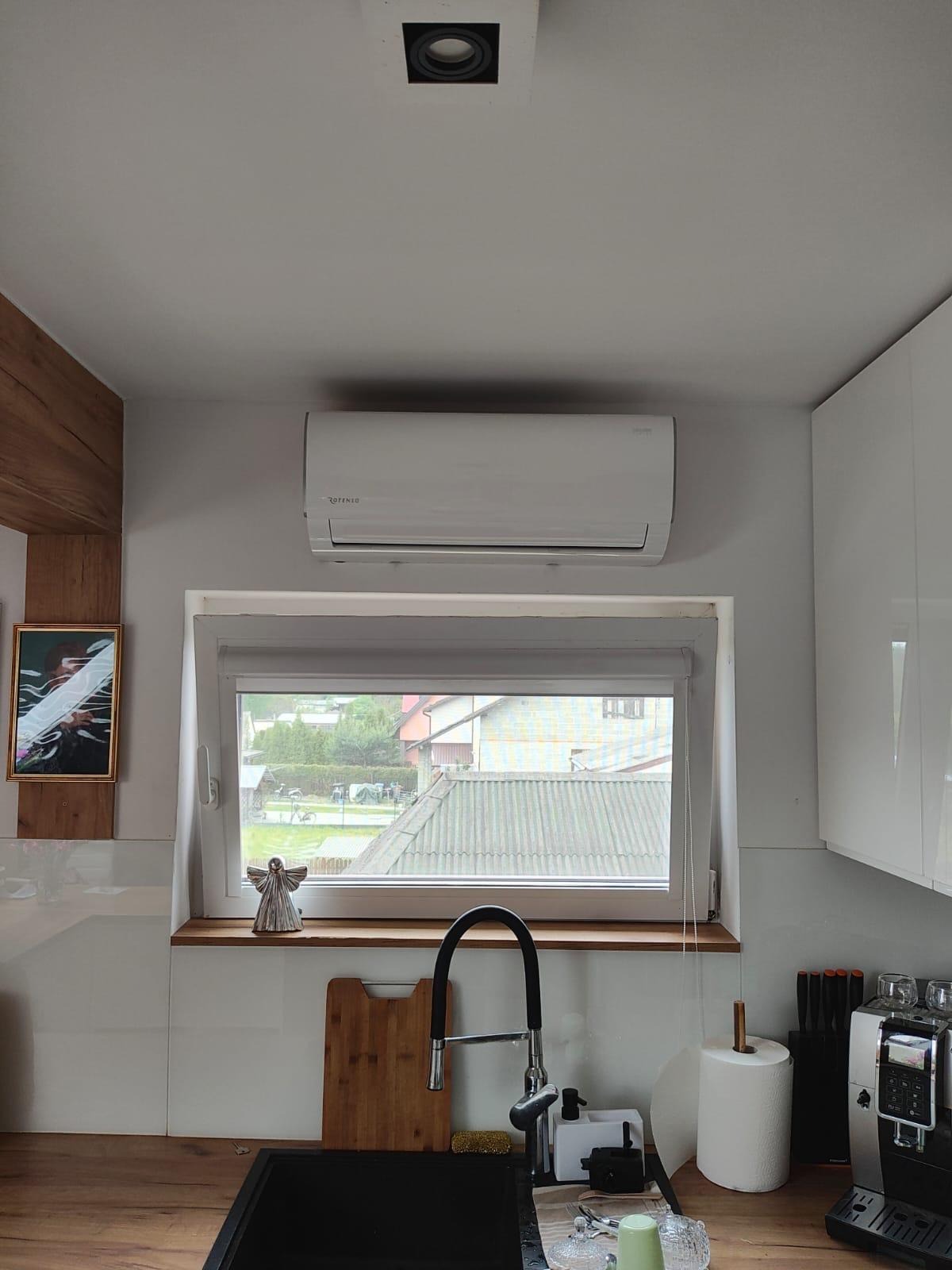 Biały klimatyzator Rotenso zamontowany nad oknem w kuchni w Końskich. Widok na zewnątrz, jasne meble kuchenne.