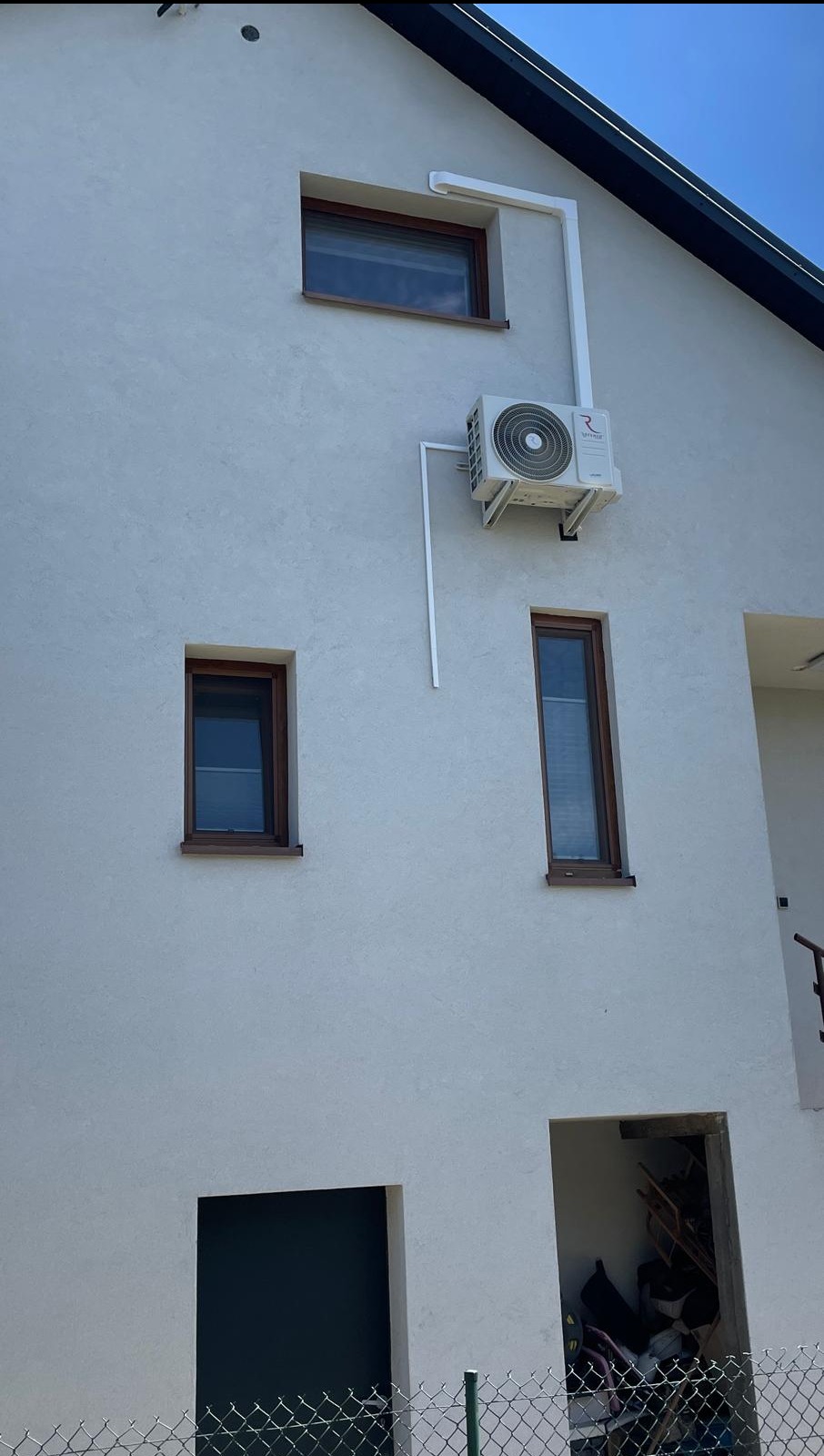 Biała elewacja domu z zamontowaną jednostką zewnętrzną klimatyzacji, widoczne okna z brązowymi ramami i białe rury odprowadzające skropliny.