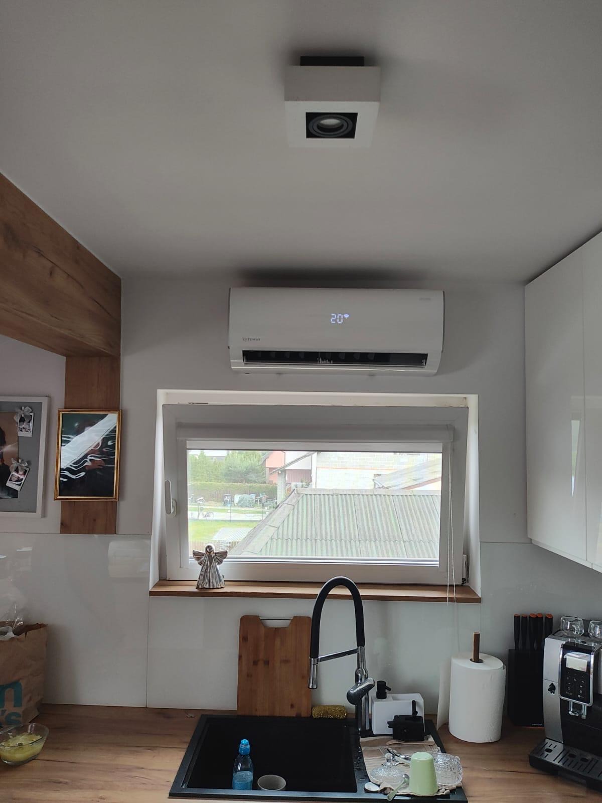 Biały klimatyzator z wyświetlaczem 20 stopni Celsjusza zamontowany nad oknem w jasnej kuchni, widok na zewnątrz z dachem budynku gospodarczego.