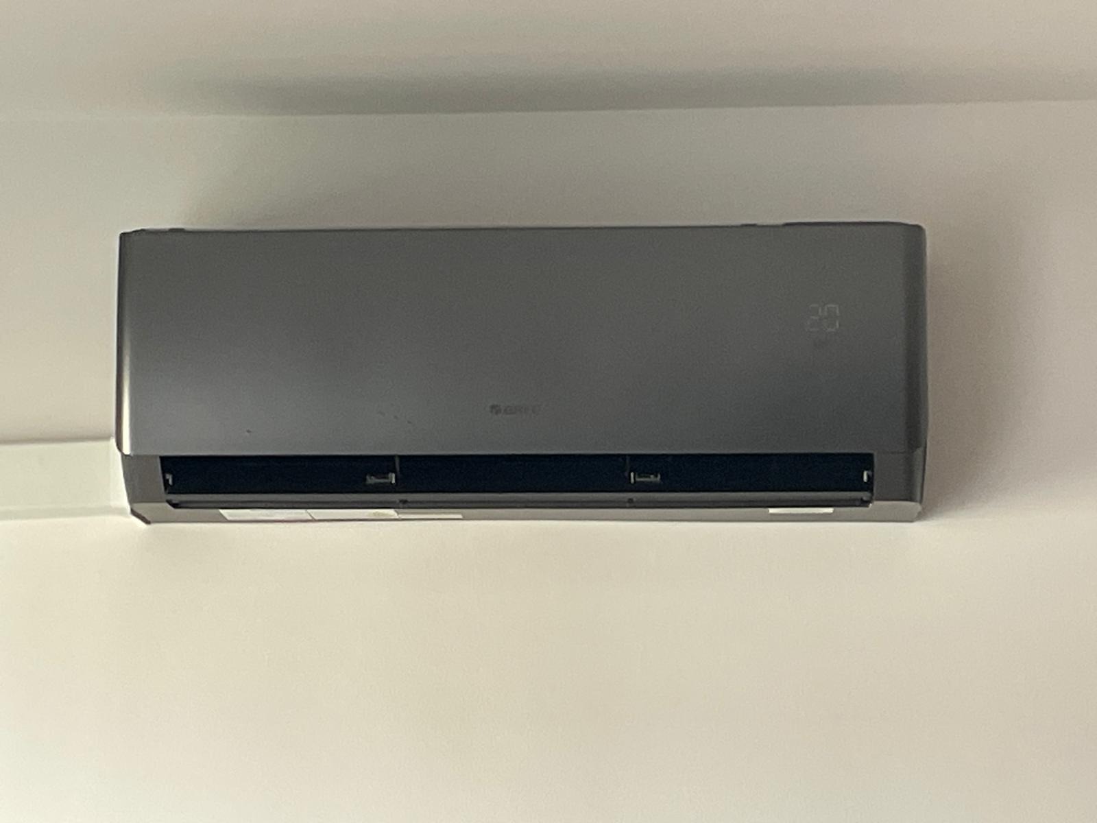 Szara jednostka wewnętrzna klimatyzacji marki Gree zamontowana na białej ścianie, wyświetlająca temperaturę 20 stopni Celsjusza.