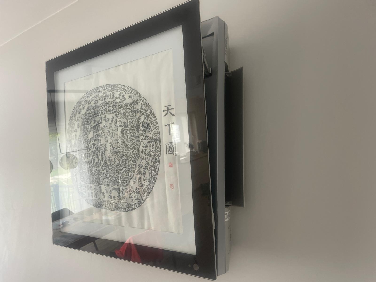 Klimatyzator ścienny z panelem dekoracyjnym w ramie, imitujący obraz z chińskimi znakami, zamontowany na jasnej ścianie.