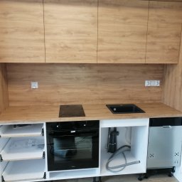 Meble na miare - Nowoczesna zabudowa kuchenna z drewnopodobnymi frontami, czarną płytą grzewczą i zlewozmywakiem, z widocznymi szufladami i sprzętem AGD w trakcie instalacji.