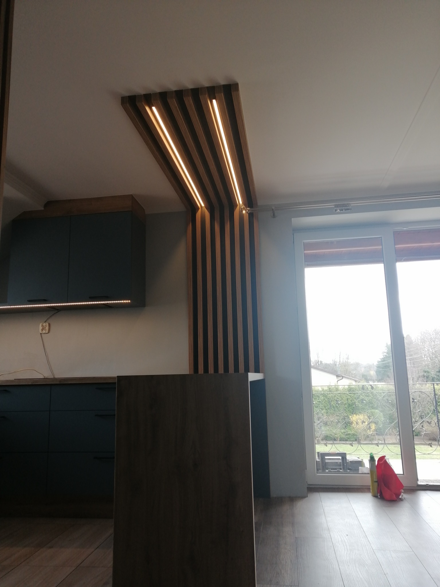 Nowoczesna kuchnia z drewnianą zabudową, oświetleniem LED w suficie i ciemnymi szafkami, widok na okno z zielonym ogrodem w tle.