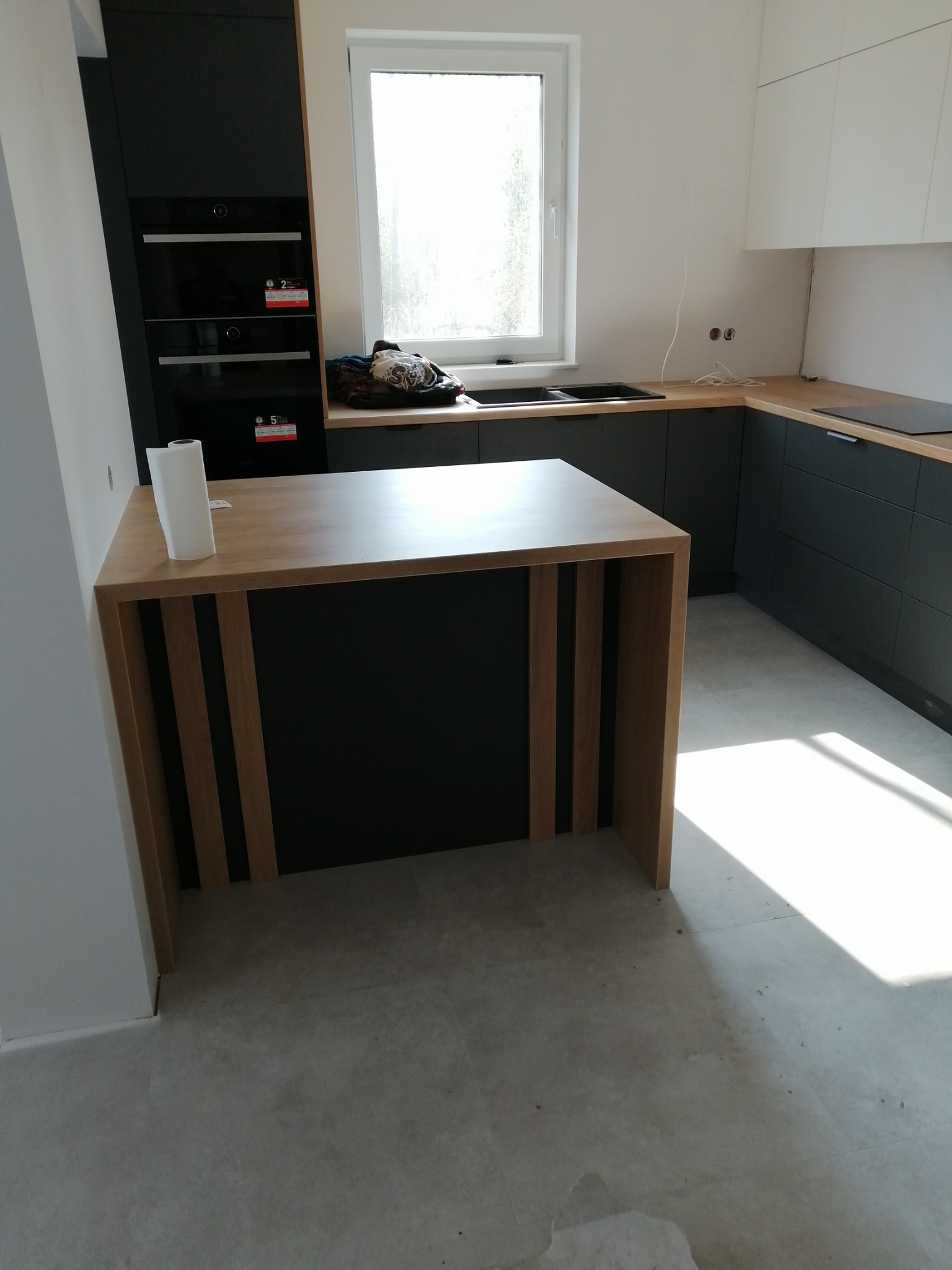 Nowoczesna kuchnia z czarnymi szafkami i drewnianym blatem wyspowym z pionowymi drewnianymi wstawkami, widoczne niedoskonałości wykończenia i niedbale położone kable elektryczne.