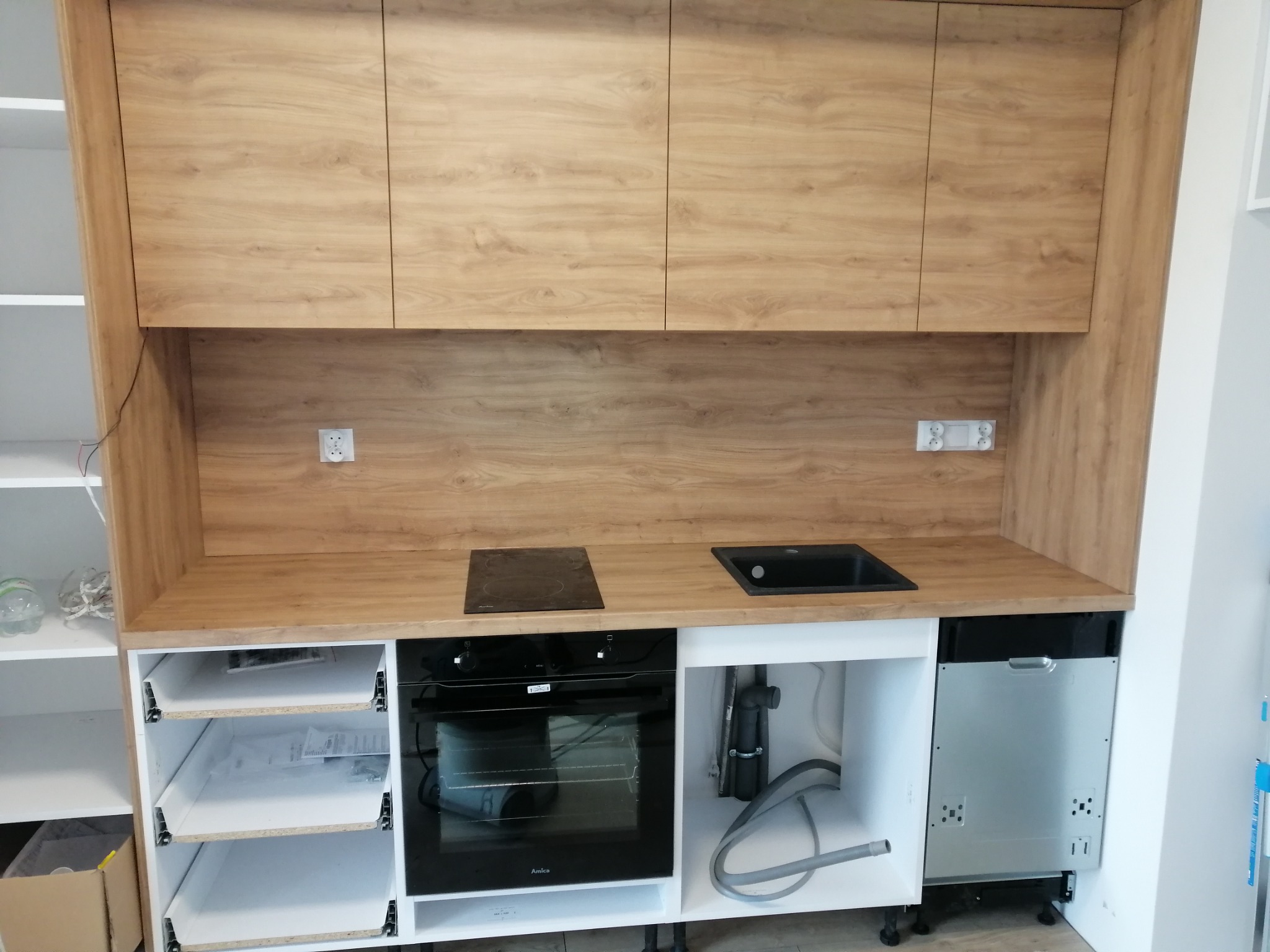 Nowoczesna zabudowa kuchenna z drewnopodobnymi frontami, czarną płytą grzewczą i zlewozmywakiem, z widocznymi szufladami i sprzętem AGD w trakcie instalacji.