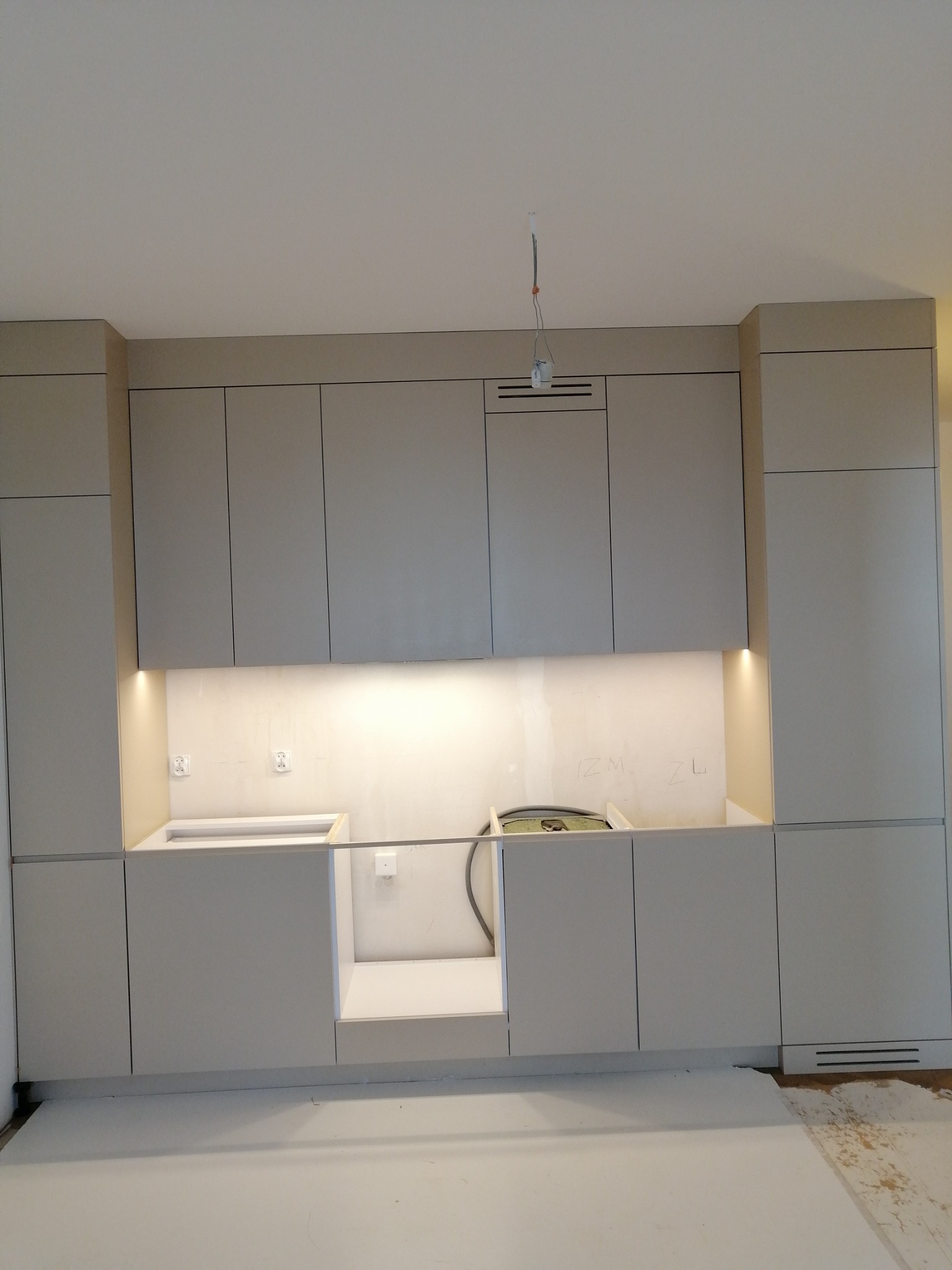 Szare szafki kuchenne w trakcie montażu, widoczne niedokończone podłączenia elektryczne i hydrauliczne, oświetlenie podszafkowe włączone, ściana z napisami.