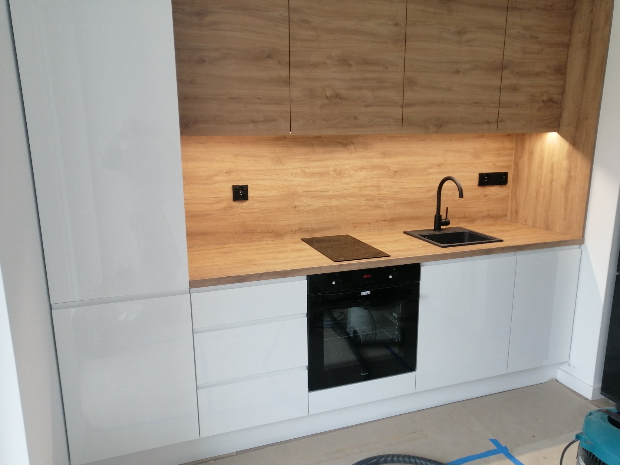 Nowoczesna kuchnia z białymi szafkami na wysoki połysk i drewnopodobnymi szafkami górnymi, czarną armaturą i płytą indukcyjną, oświetleniem podszafkowym, częściowo widoczny odkurzacz na podłodze.