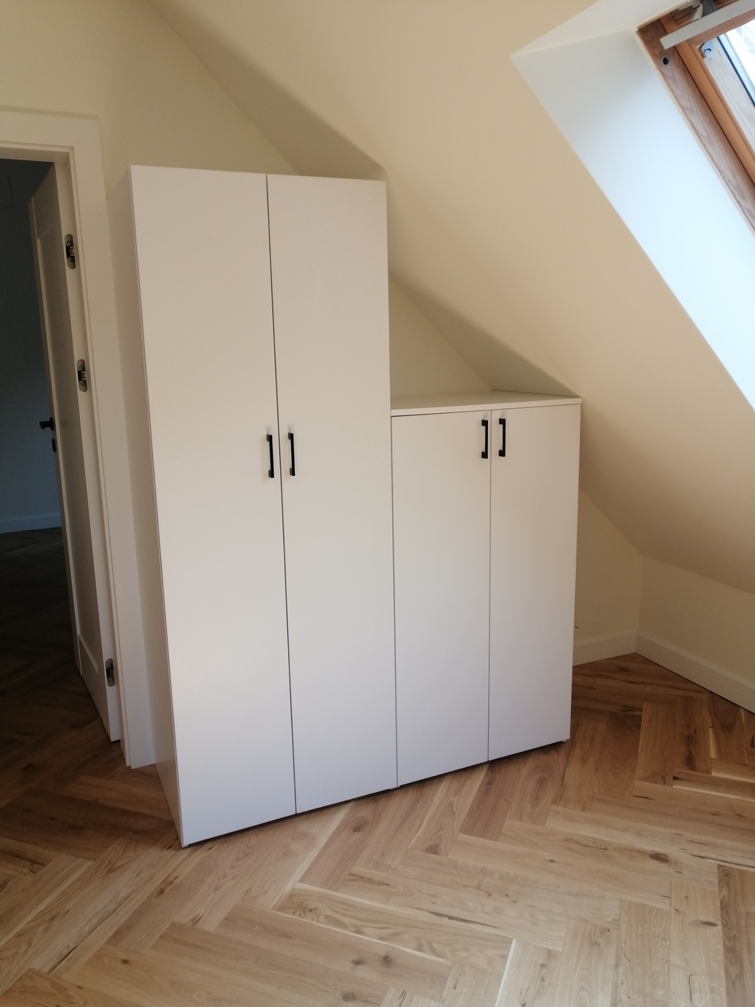 Białe szafy z czarnymi uchwytami, dopasowane do skosu poddasza, na parkiecie ułożonym w jodełkę.