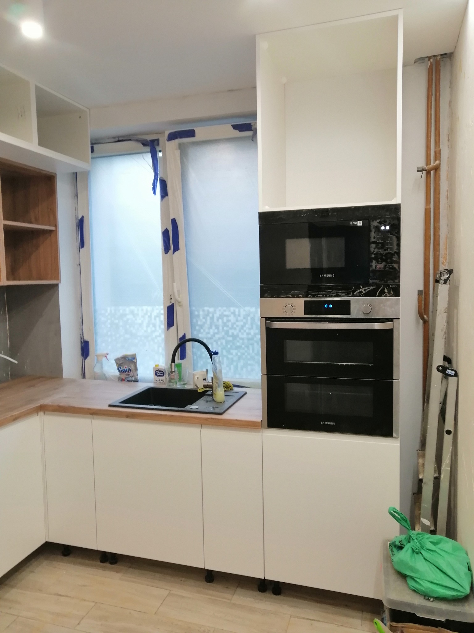 Wnętrze kuchni w trakcie montażu: białe szafki z drewnianym blatem, czarny zlew, okno z niebieską taśmą malarską, wbudowany piekarnik i mikrofalówka marki Samsung, drabina oparta o ścianę, podłoga...