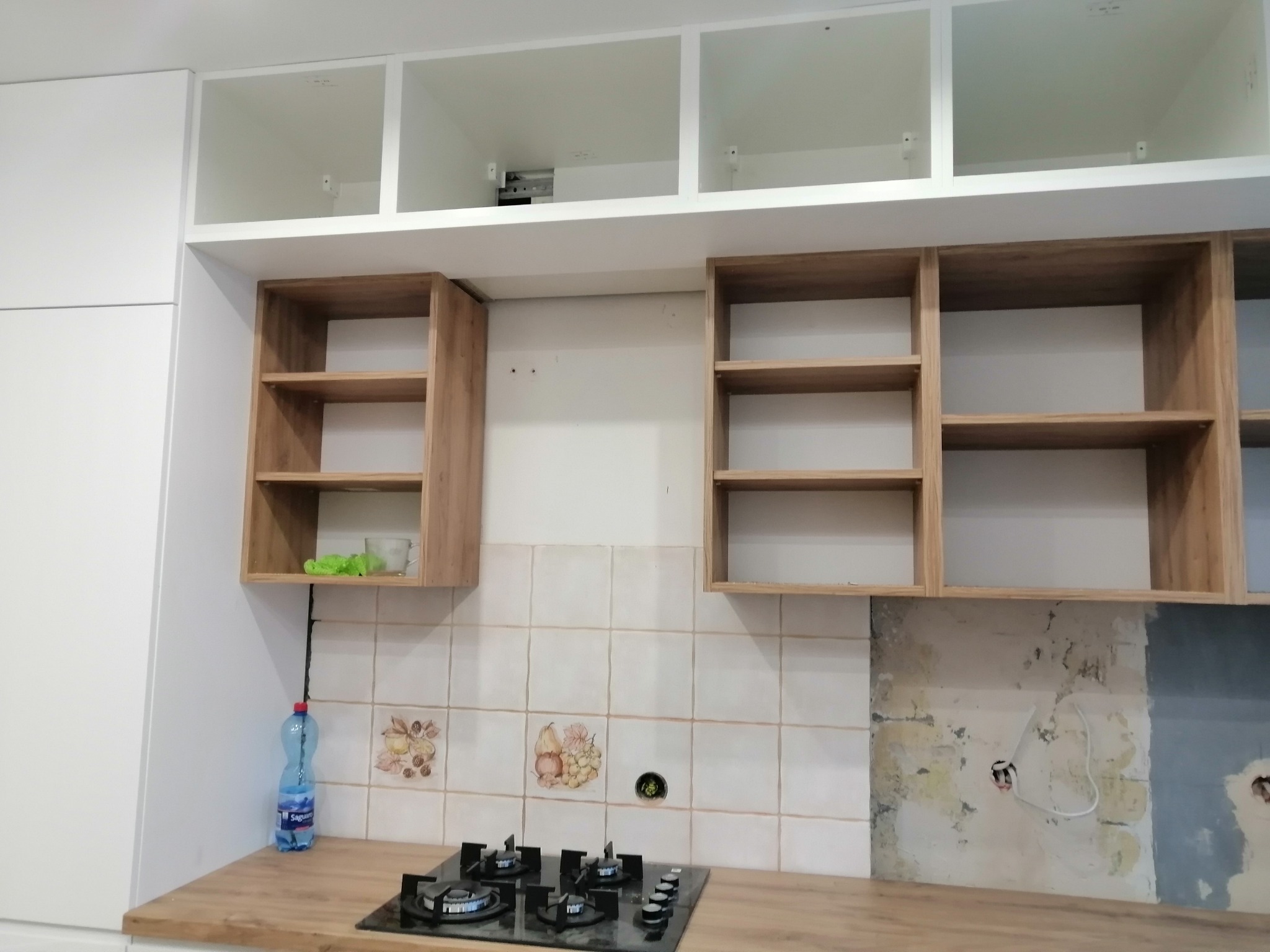 Częściowo zamontowane szafki kuchenne w trakcie remontu: białe szafki górne i drewniane półki wiszące nad kuchenką gazową, widoczne niedokończone płytki ścienne.