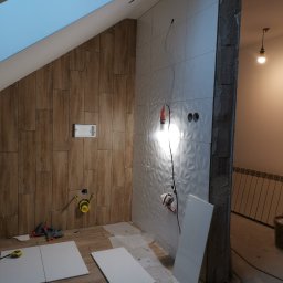 Mar-Dom Marek Adamczyk - Wnętrze łazienki w trakcie remontu: częściowo ułożone drewnopodobne i białe płytki ścienne z wzorem 3D, widoczne narzędzia, przewody elektryczne i fragment sufitu skośnego z oknem dachowym.