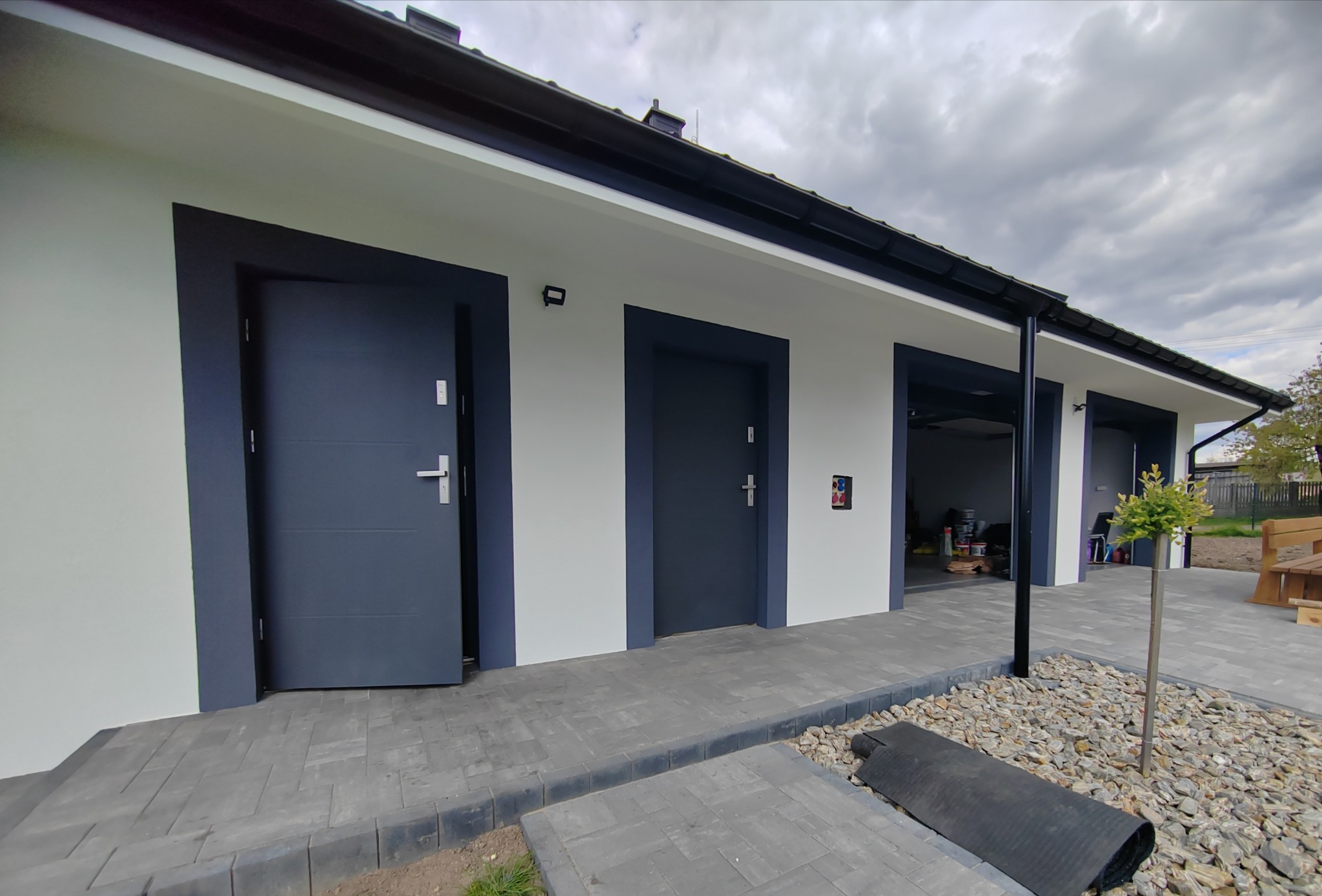 Nowoczesny budynek z trzema ciemnoszarymi drzwiami, białą elewacją i ciemnym obramowaniem, z widocznymi elementami wykończeniowymi oraz fragmentem brukowanej ścieżki i kamienistego ogrodu.