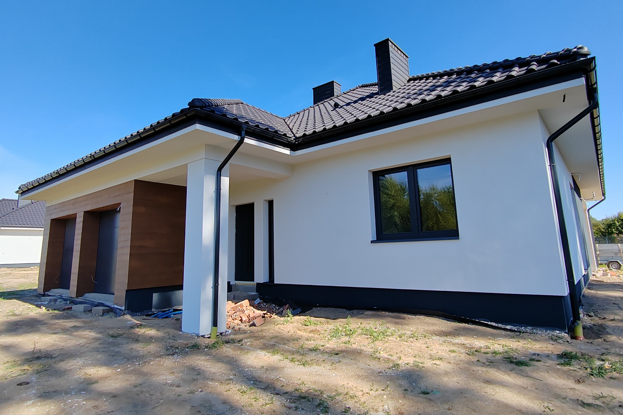 Nowoczesny dom jednorodzinny z białą elewacją, czarną stolarką okienną i grafitową dachówką, w trakcie prac wykończeniowych na zewnątrz, widoczne materiały budowlane.