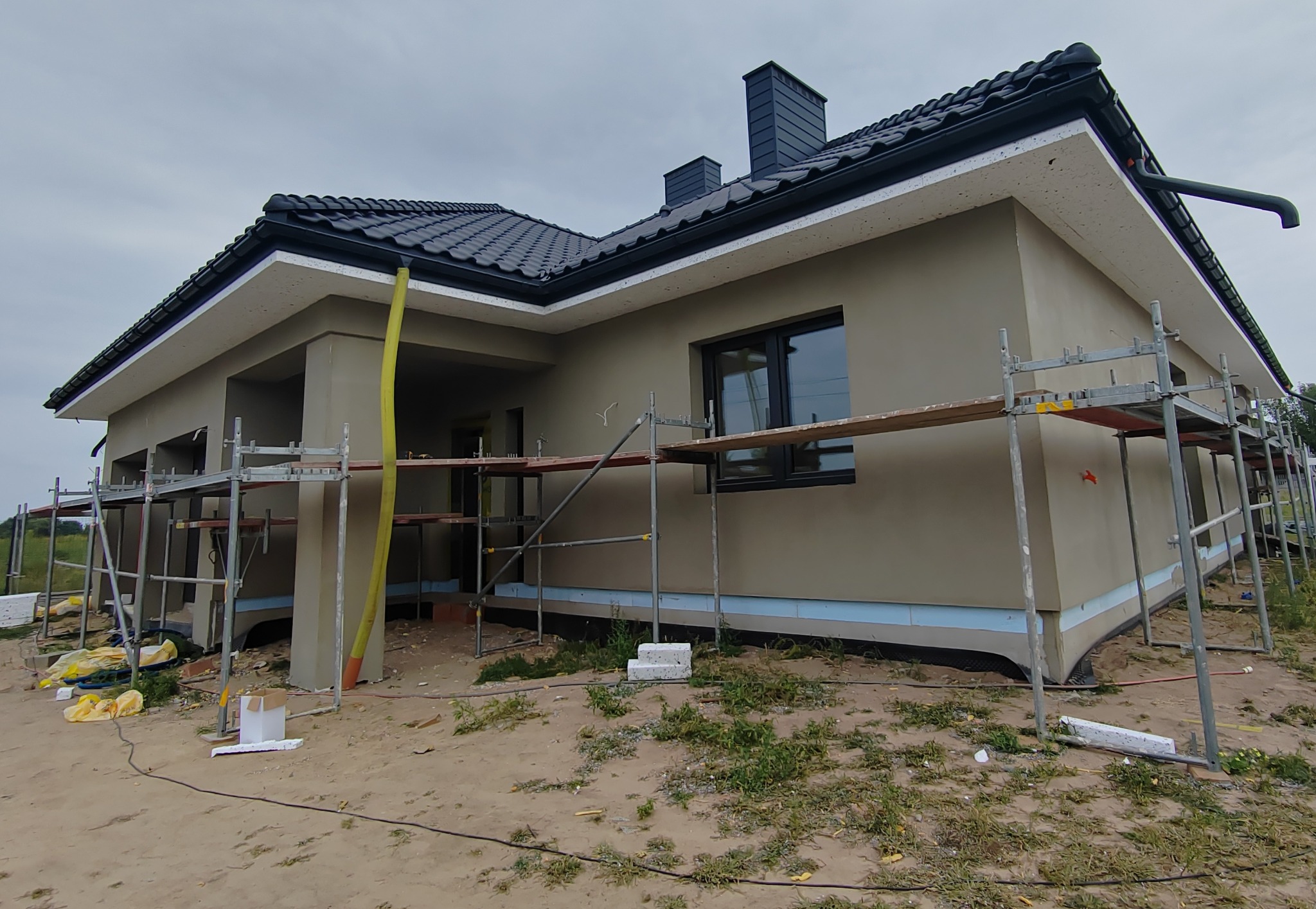 Dom jednorodzinny w trakcie ocieplania elewacji, widoczne rusztowania, ocieplenie styropianem i nałożony tynk, dachówka ceramiczna w kolorze grafitowym.
