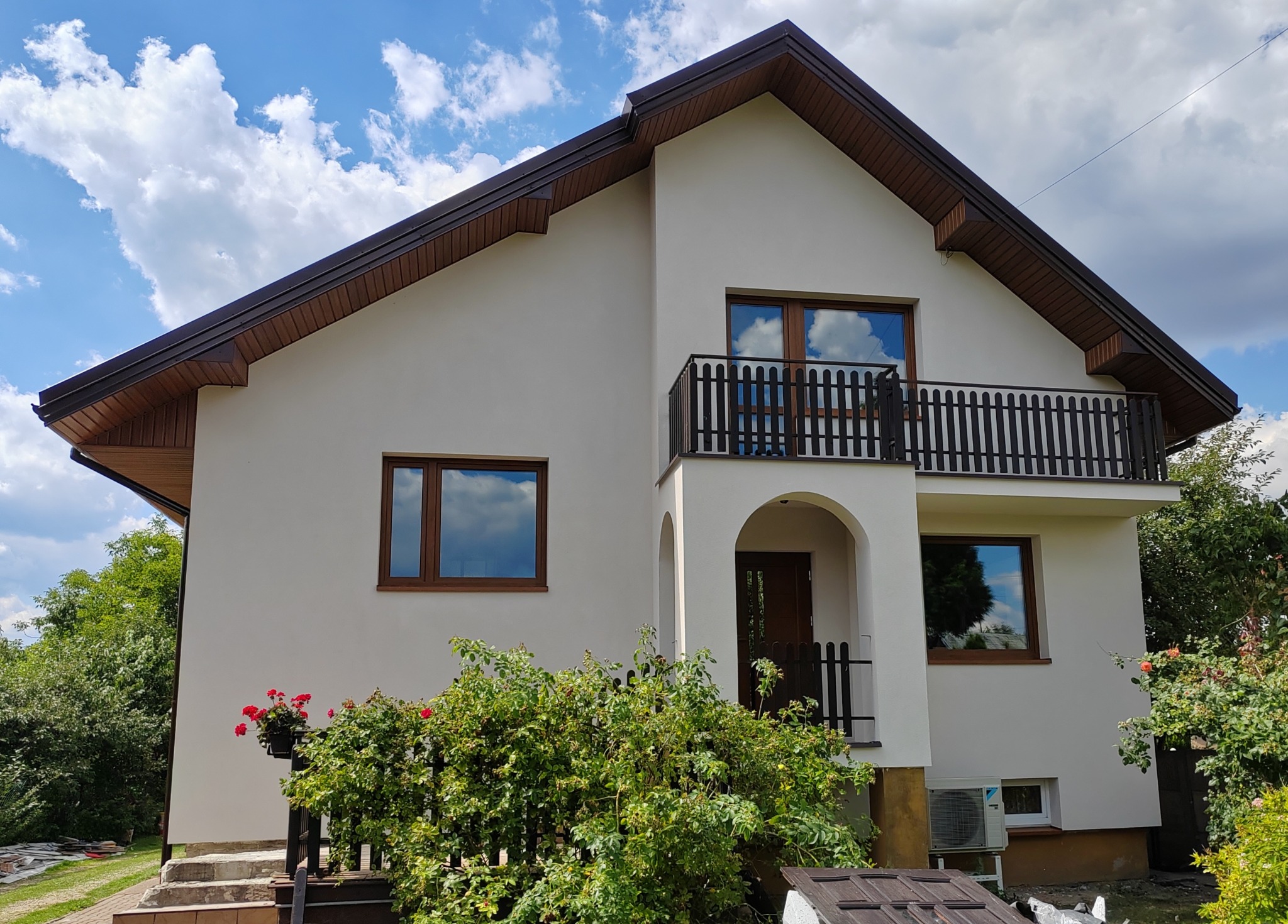 Jasny, dwukondygnacyjny dom z balkonem w Opocznie po pracach elewacyjnych, z widocznymi drewnianymi oknami i brązowym dachem na tle błękitnego nieba z chmurami.
