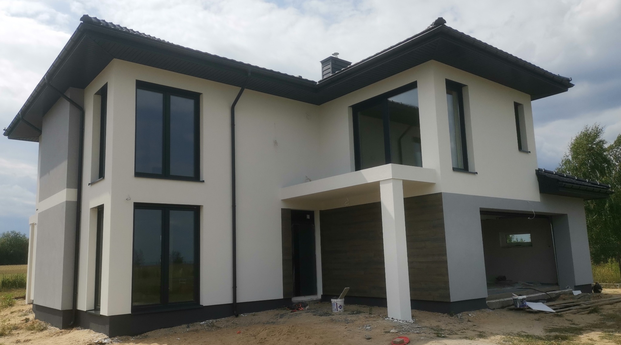 Dwukondygnacyjny dom z płaskim dachem, z fragmentami elewacji w odcieniach szarości i bieli, widoczne narzędzia malarskie i wiadro z farbą przy wejściu.