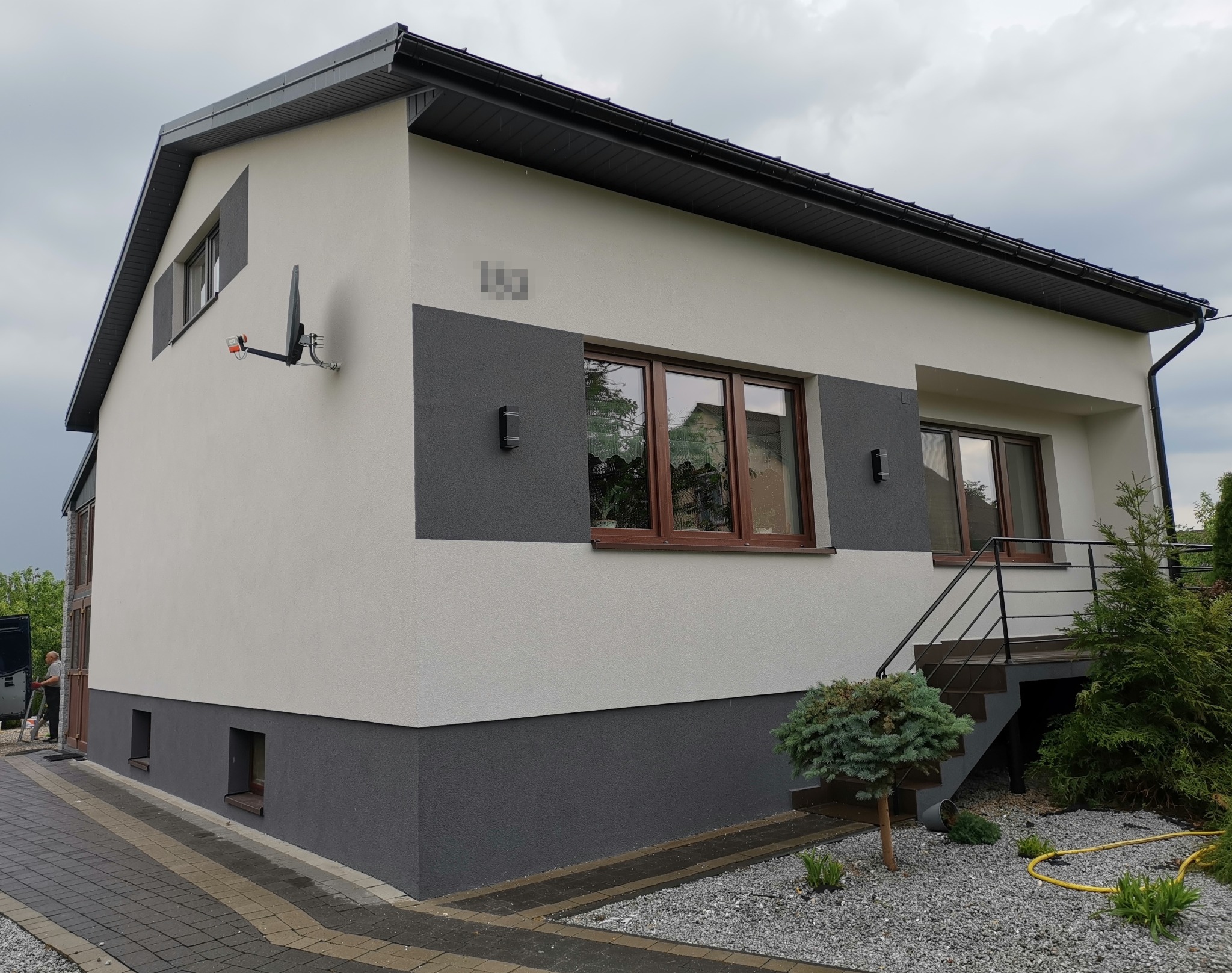 Dwukondygnacyjny dom z elewacją w odcieniach szarości i bieli, z widoczną anteną satelitarną, oknami z brązowymi ramami, zewnętrznymi lampami i schodami wejściowymi z czarną balustradą.