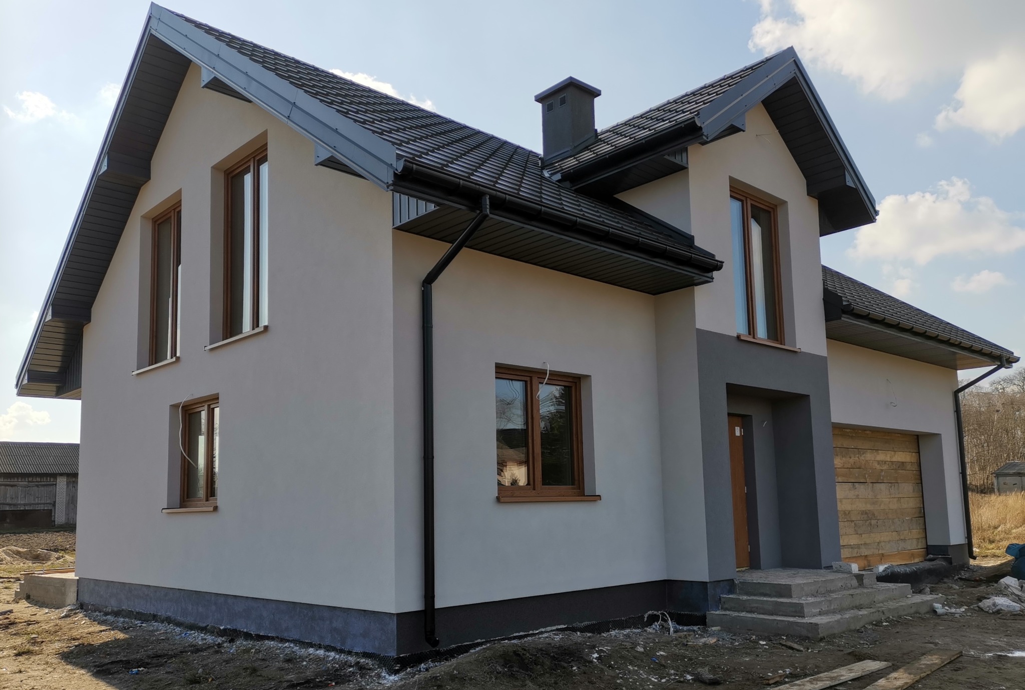 Nowo wykończony dom jednorodzinny z szarą elewacją, brązowymi oknami i ciemnoszarą dachówką, widoczne elementy orynnowania i fragment terenu przed budynkiem z resztkami materiałów budowlanych.