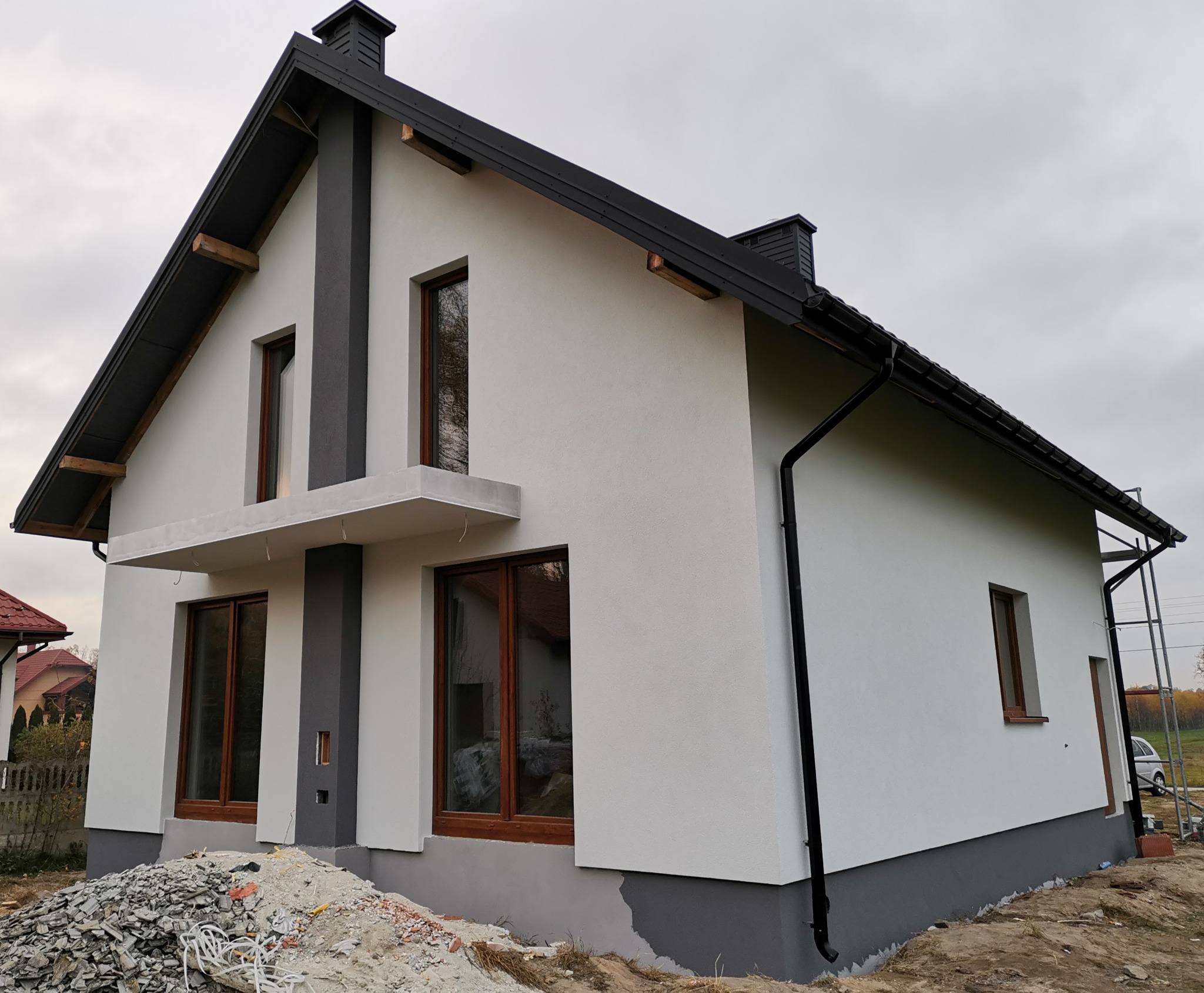 Nowoczesny dom jednorodzinny z białą elewacją i grafitowymi elementami dekoracyjnymi, widoczne okna z drewnianą stolarką, fragment dachu z czarną blachodachówką, przy ziemi pozostałości gruzu.